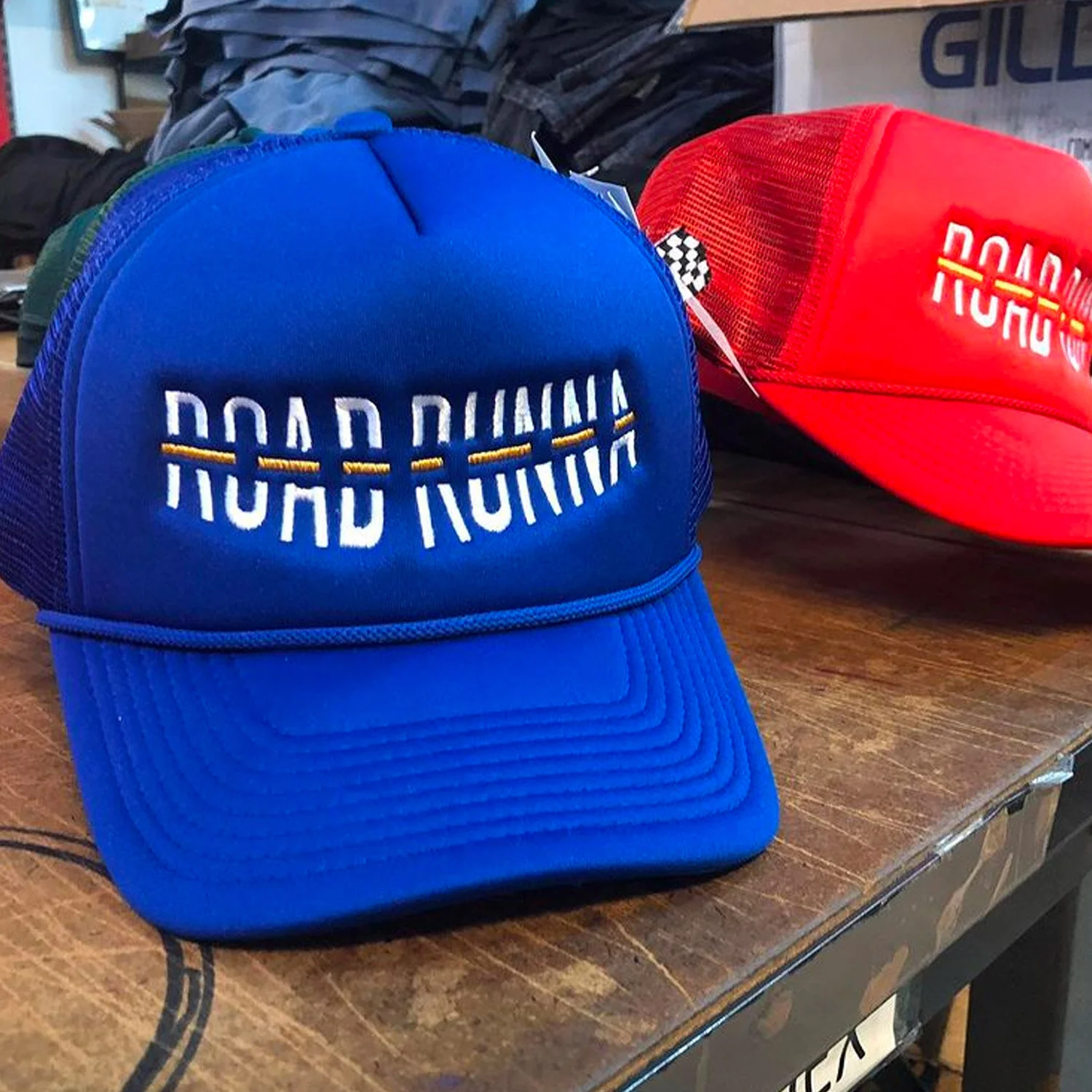 A blue trucker style baseball cap with the words 'RACE DAY' embroidered on the front, placed on a wooden surface. A red cap with a similar design is in the background.