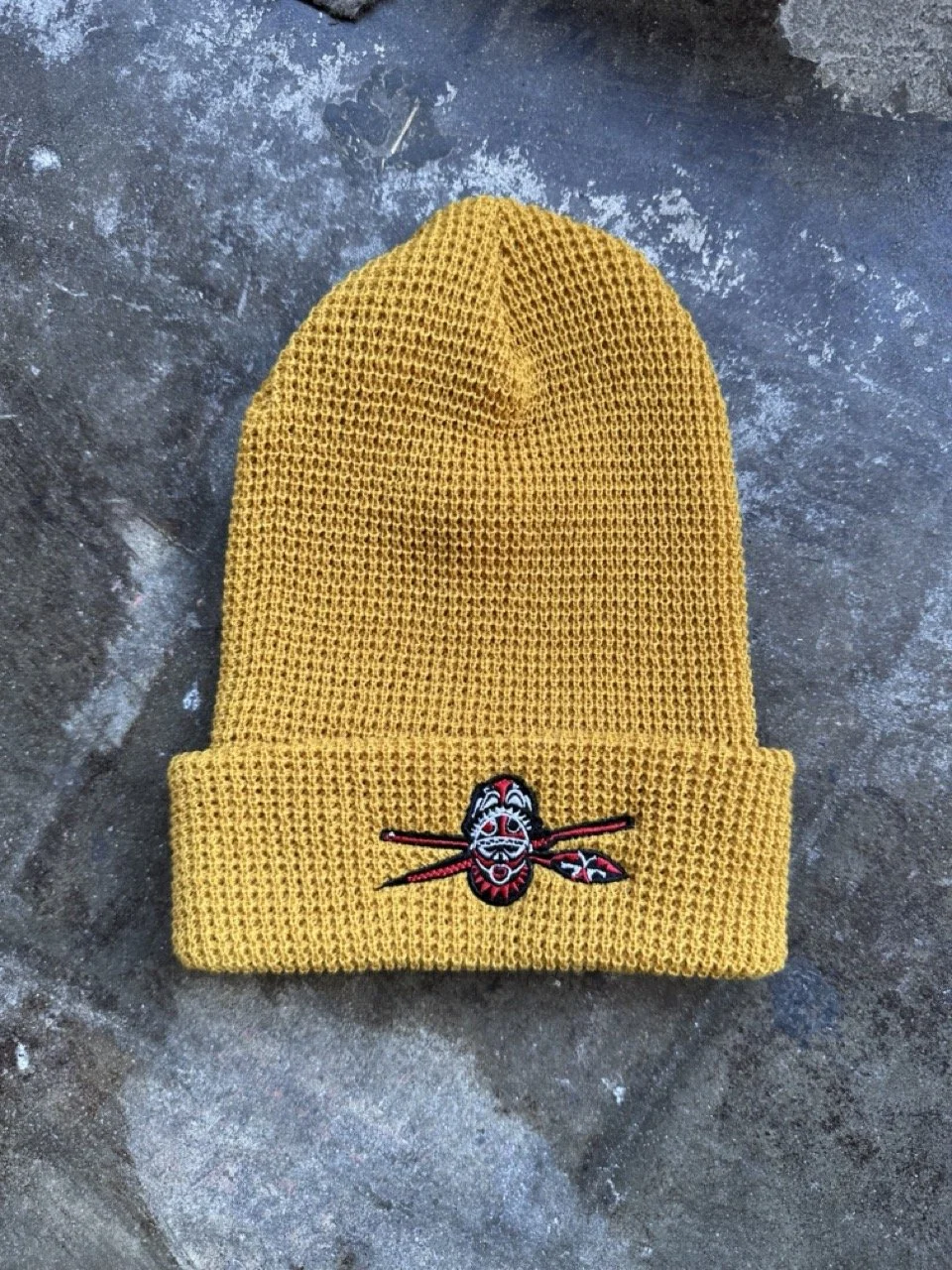 Yellow knit beanie hat with a colorful embroidered face and spear design, lying on a dark, textured concrete surface.