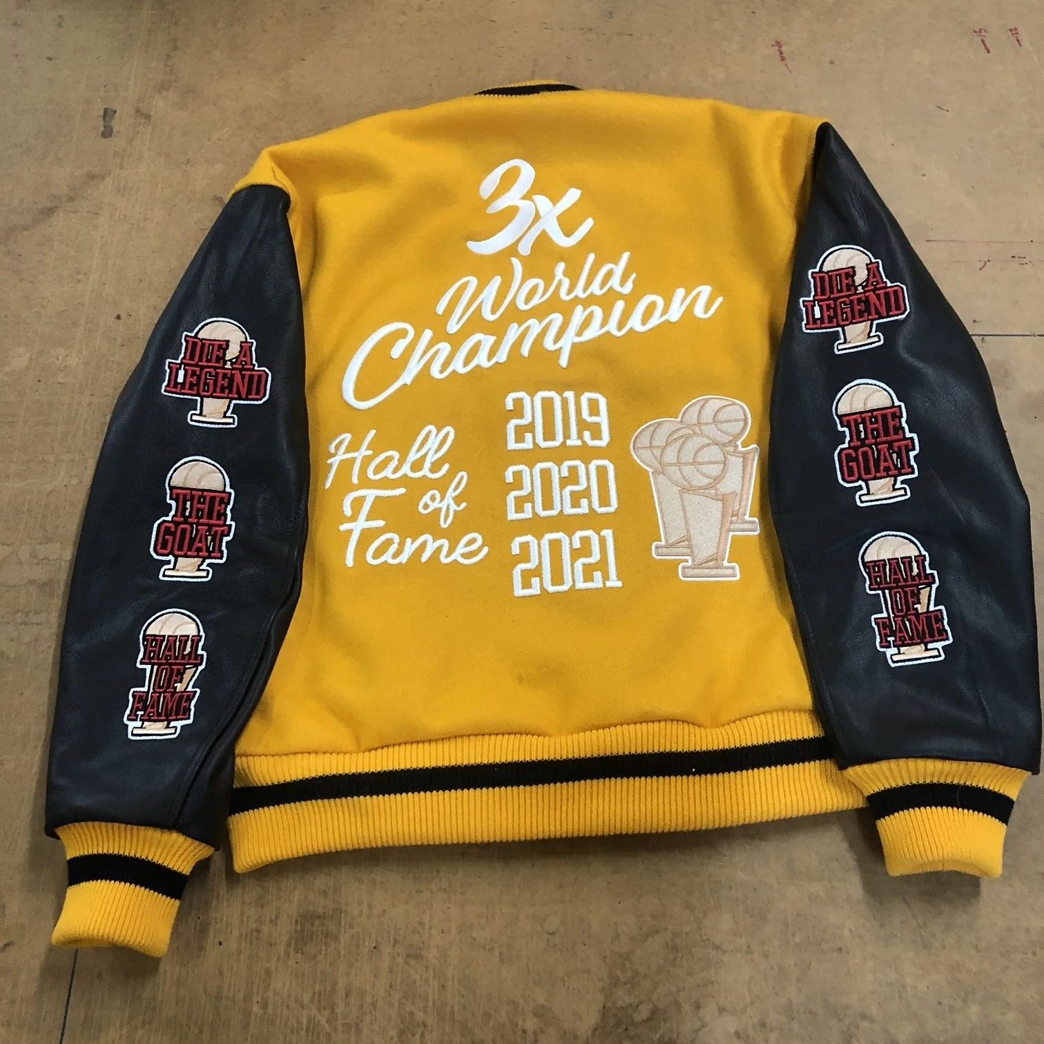 Yellow and black varsity jacket with patches and embroidery celebrating a basketball team's championship wins from 2019 to 2021, featuring a trophy graphic.