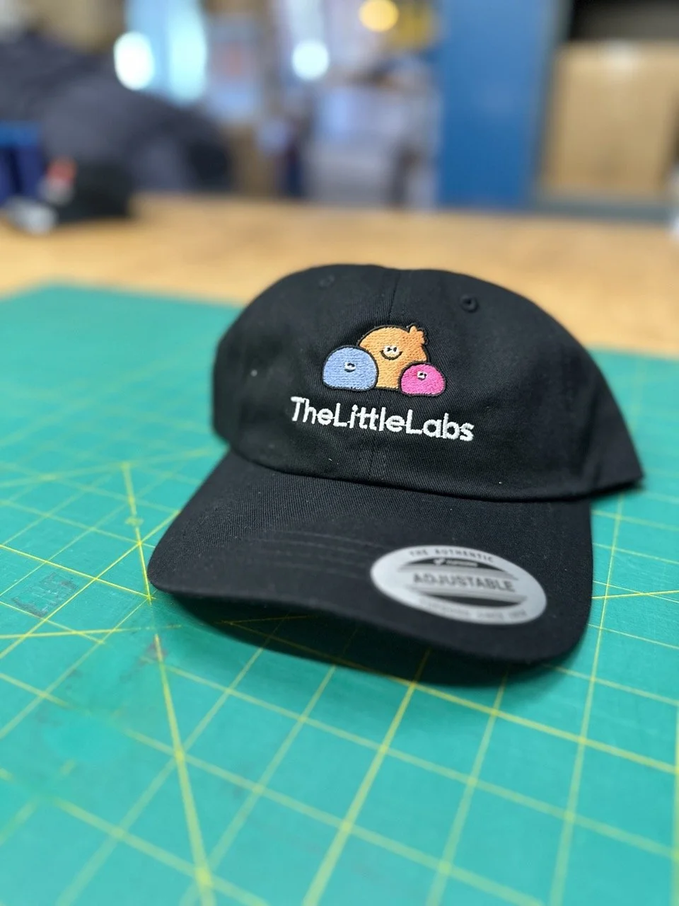 Black adjustable cap with embroidered logo of three cute, smiling bacteria characters in blue, orange, and pink, and the text 'TheLittleLabs' underneath on a green patterned cutting mat.