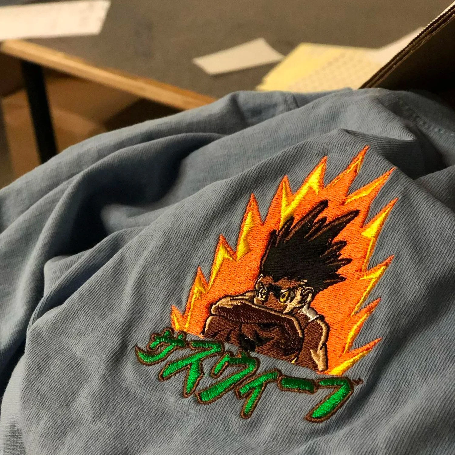 Close-up of an embroidered patch on a gray fabric, depicting a character with spiky black hair, intense eyes, and a fighting stance, surrounded by orange and yellow flames and green Japanese characters below.