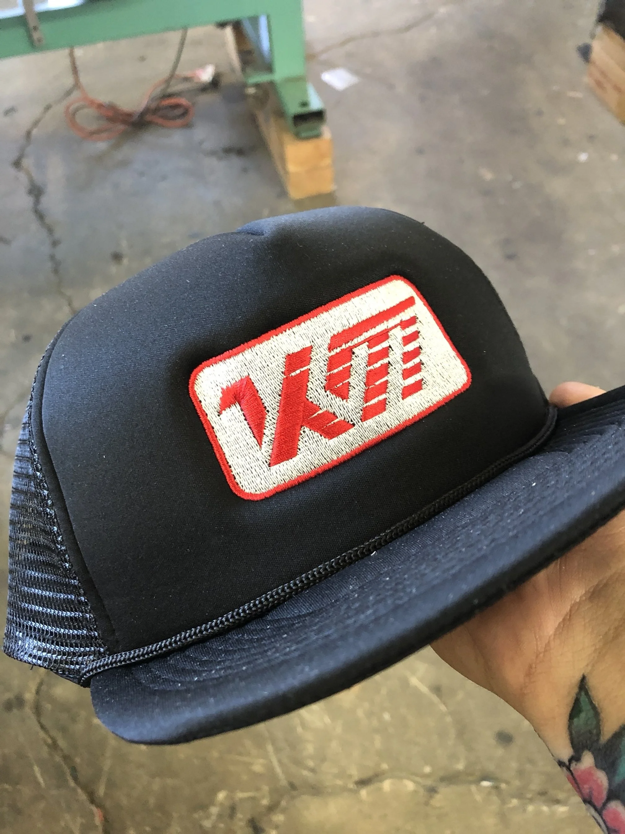 Close-up of a black Velo trucker cap with red and white embroidered Velo logo, held by a hand with a tattoo, in a workshop environment.