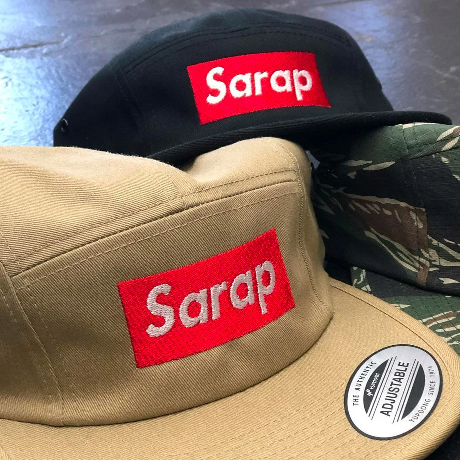Two baseball caps, one black and one beige, both with red and white "Sarap" logos, and a camouflage-patterned hat.