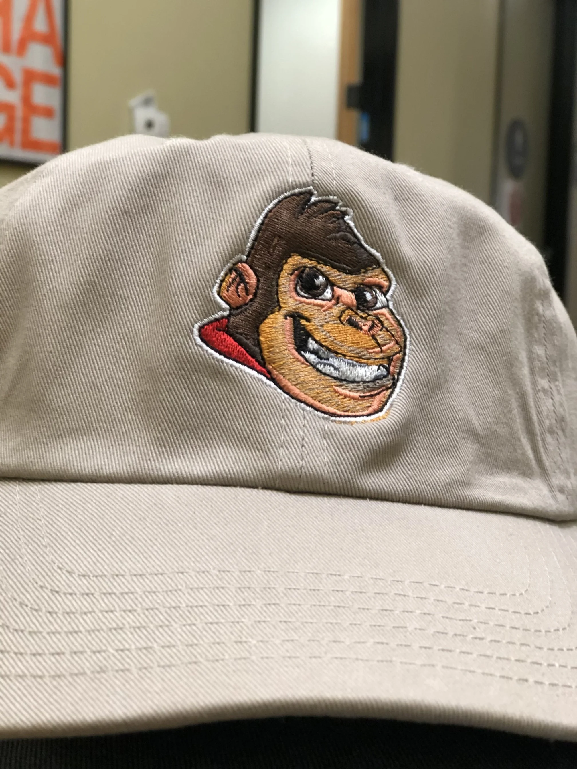 A khaki baseball cap with an embroidered cartoon gorilla face smiling, with a red background behind its head.