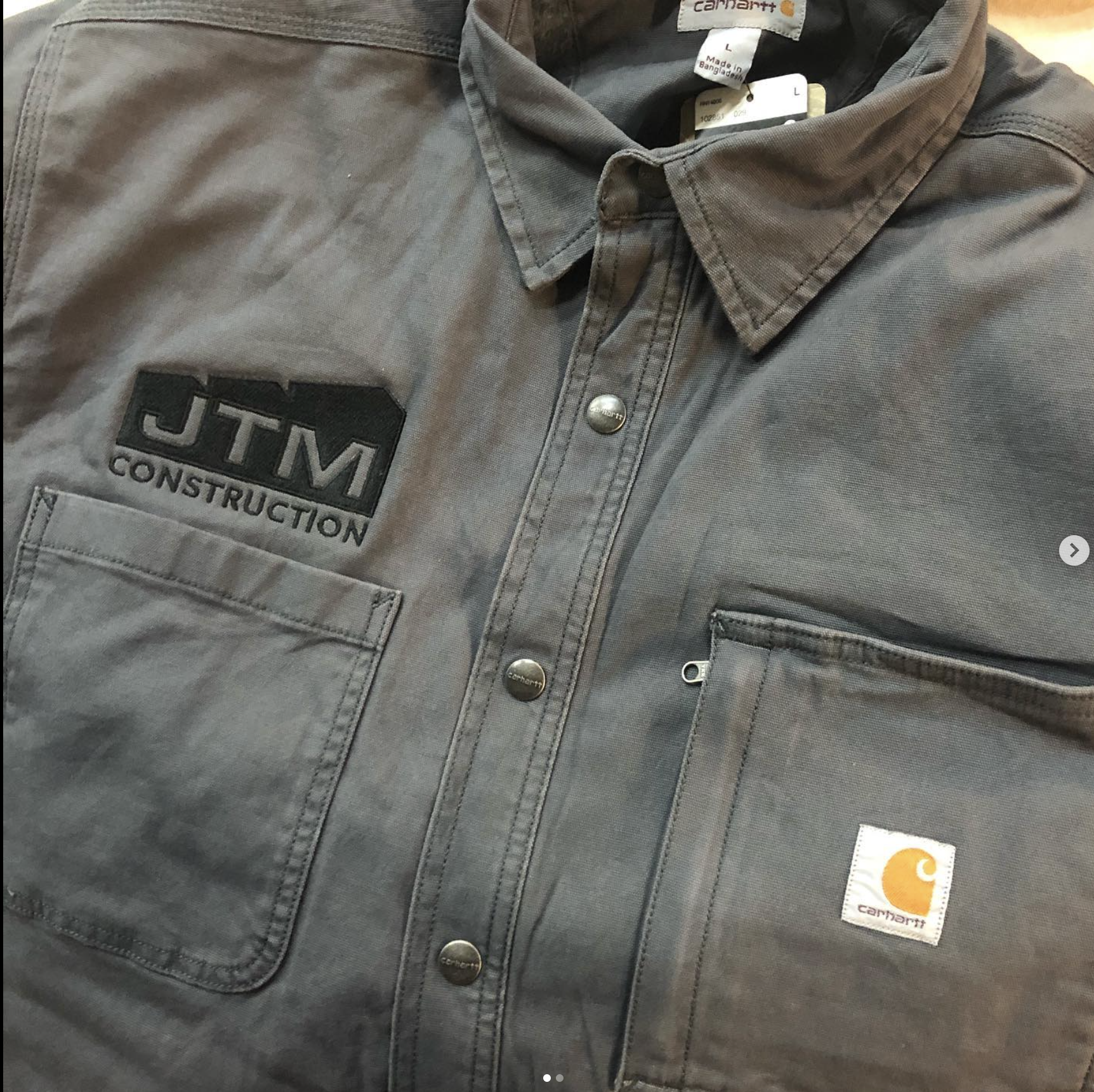 Gray Carhartt work shirt with two chest pockets, black embroidered patch reading "JTM CONSTRUCTION," and Carhartt logo tag on the right pocket.
