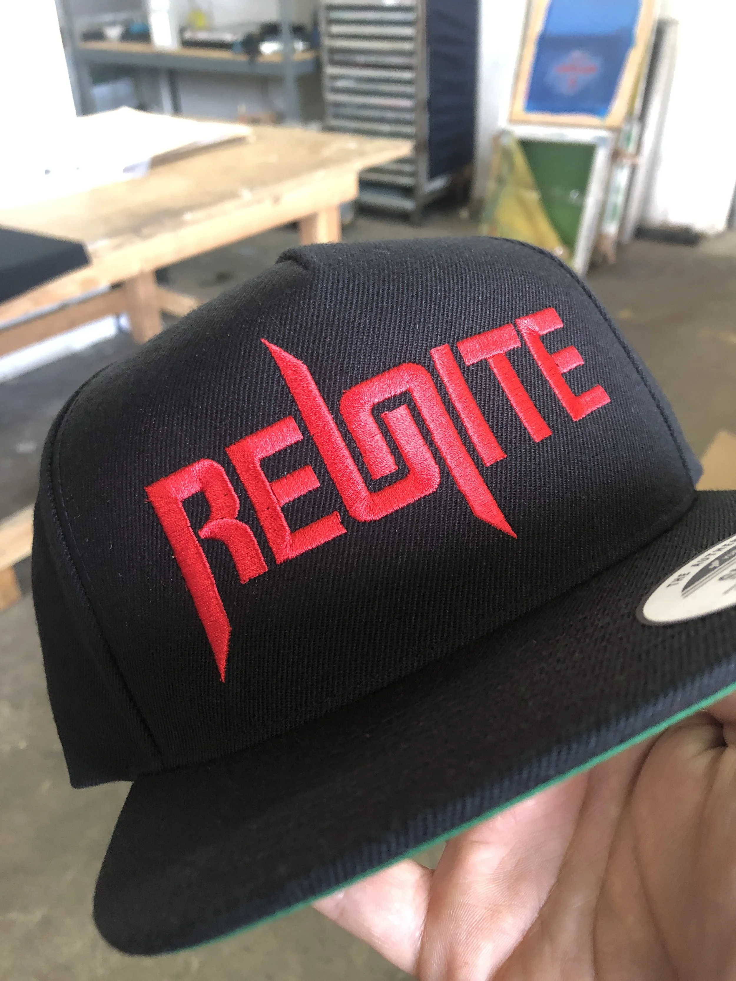 Close-up of a black cap with red embroidered text that reads 'REDDITE' in a stylized font, held by a hand in a workshop or garage setting.