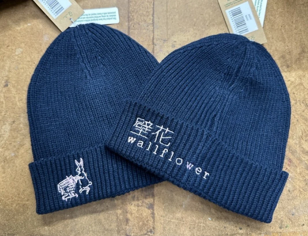 Two black knit beanies, one with a small white and purple embroidered rabbit logo on the cuff and the other with white embroidered Chinese characters and the word "wallflower" on the front, resting on a wooden surface with some paper tags in the back