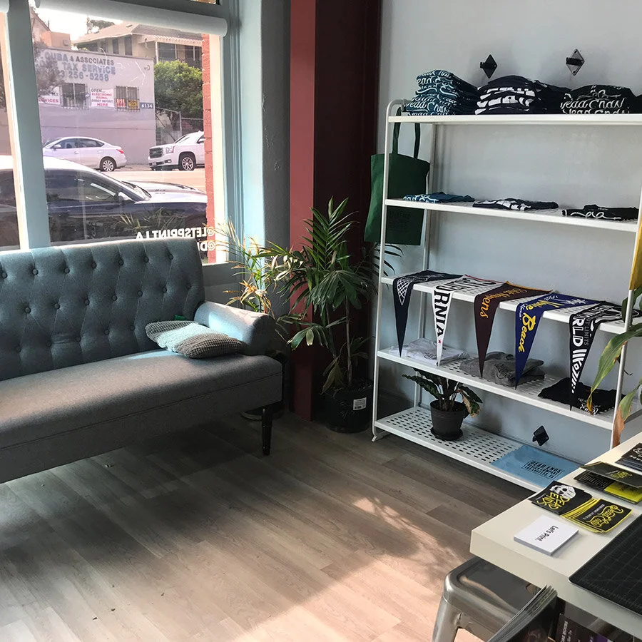 Interior of a store with a grey tufted sofa, a potted plant next to a large window, and a white shelving unit displaying various scarves, with additional items on a nearby table and a dark wood floor.