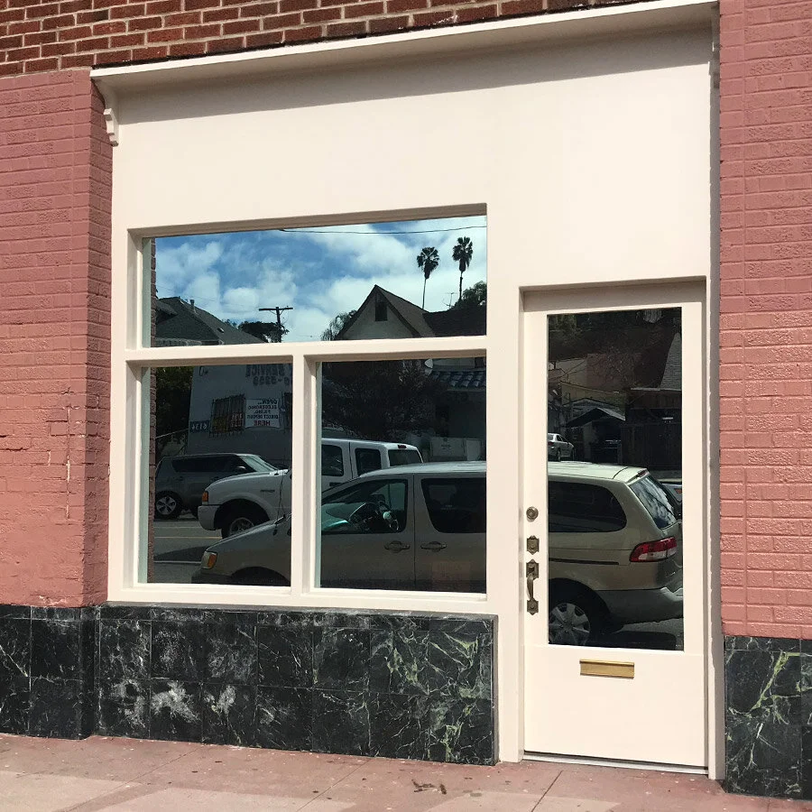 A storefront with a large glass window and a white door with a glass panel. The building has pink brick walls and a black marble base. Reflections of parked cars and houses with palm trees and a cloudy blue sky are visible in the window.