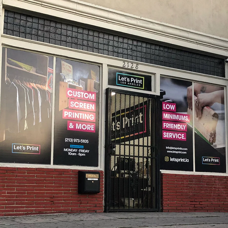 The storefront of Let's Print Los Angeles advertising custom screen printing, low minimums, friendly service, and business hours from 10 am to 6 pm, with a hanging gate in front, and sidewalk in front of the entrance.