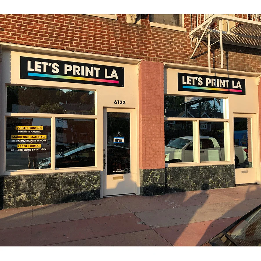 Exterior of a printing shop called 'Let's Print LA' with signage, address 6133, an 'Open' sign on the door, and window decals listing services like screen printing, marketing materials, and banner printing. The storefront features large windows and b