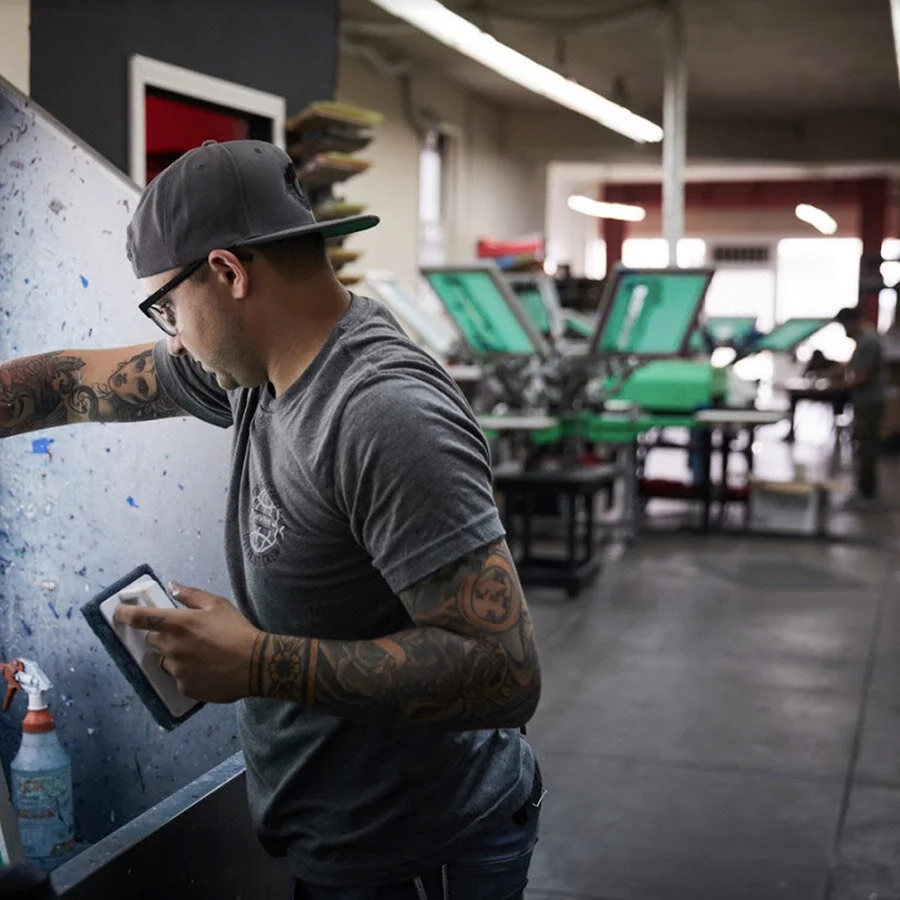 A man with tattoos, glasses, a gray t-shirt, and a backward cap is working in a print shop, operating a printing press or machine.