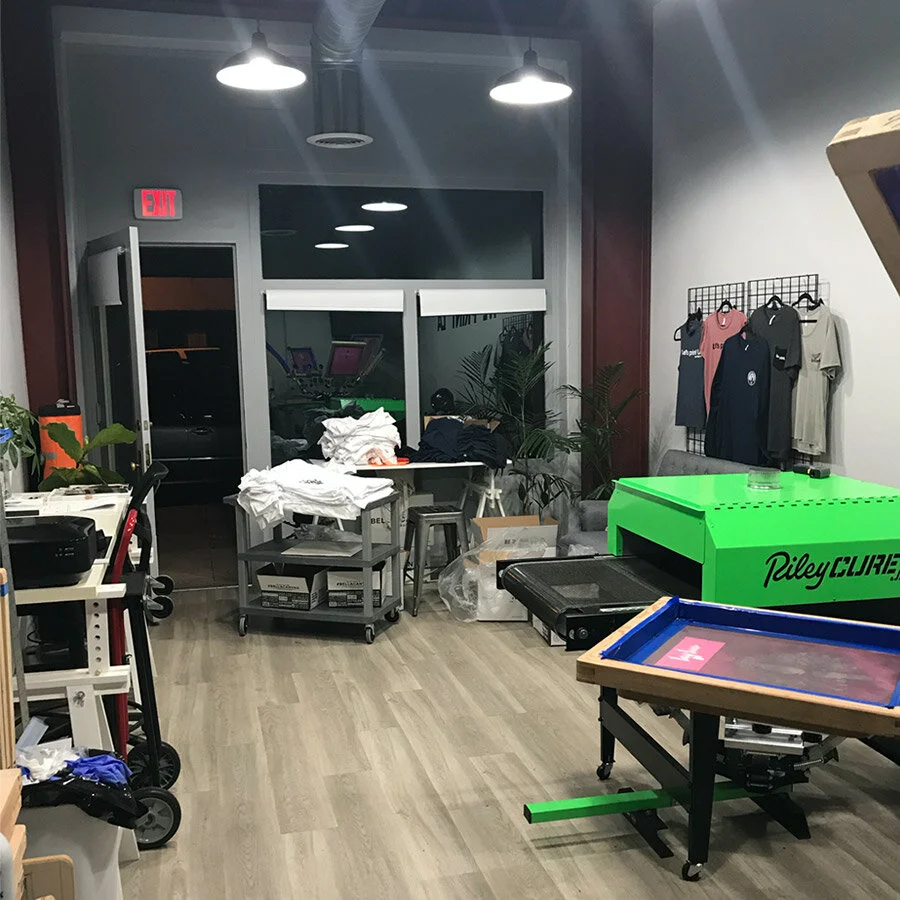 Interior of a retail or office space with merchandise, storage boxes, a pinball machine, and clothing on display, illuminated by ceiling lights.