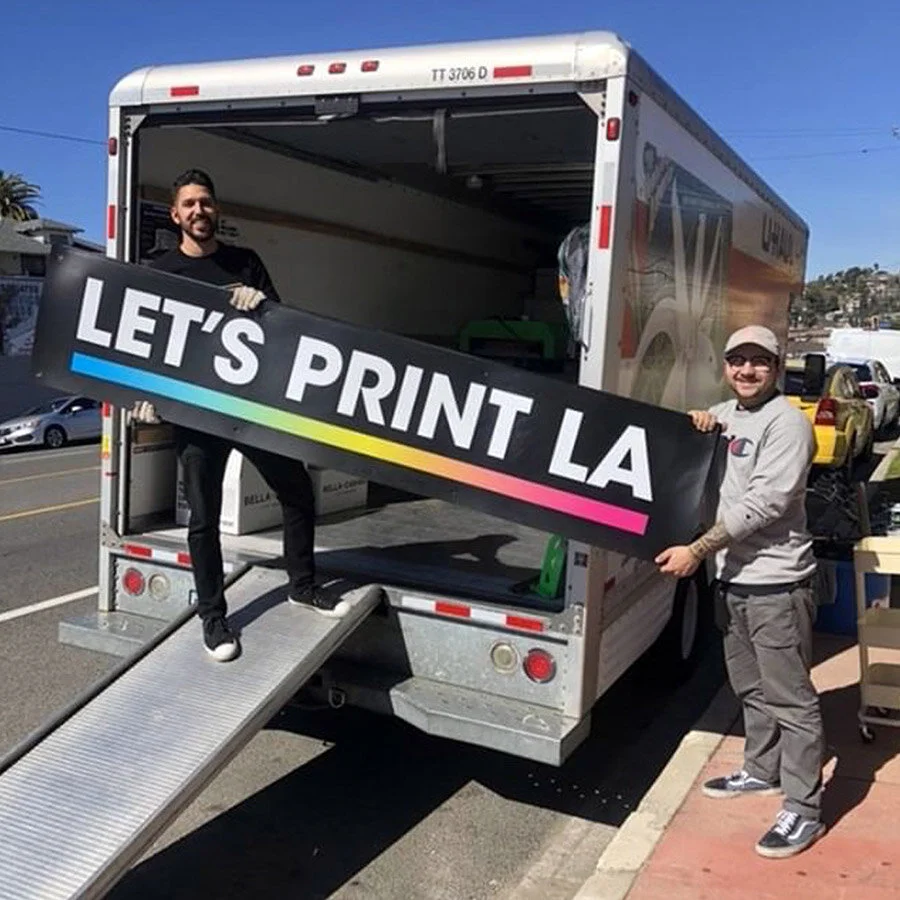Two men holding a large sign that says 'LET'S PRINT LA' in front of an open delivery truck on a sunny day.