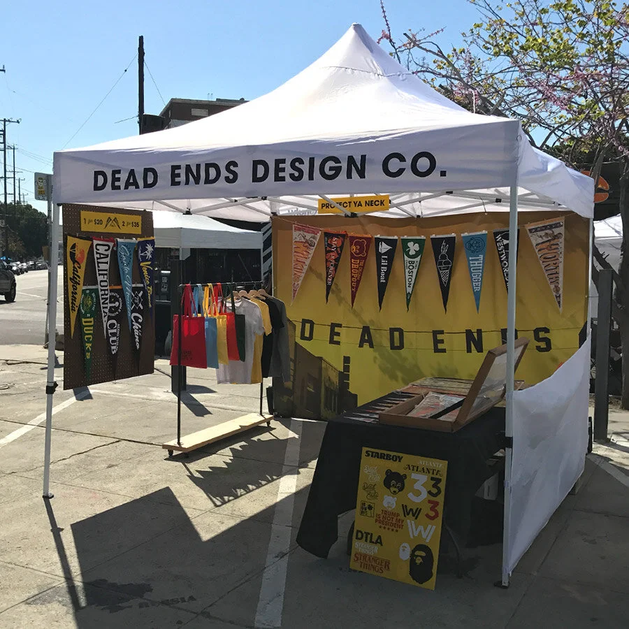 A market stall with a white canopy labeled Dead Ends Design Co. displaying colorful pennant banners, t-shirts, and posters, set up outdoors on a sidewalk.