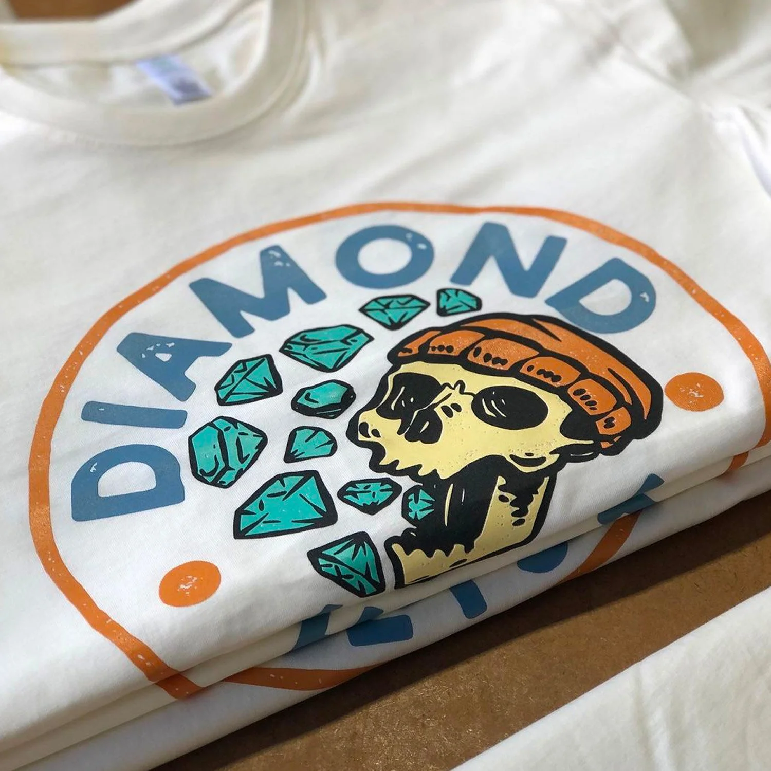 Close-up of a white t-shirt with a graphic of a skull wearing a bandana and surrounded by turquoise diamonds, with the word 'DIAMOND' partially visible.
