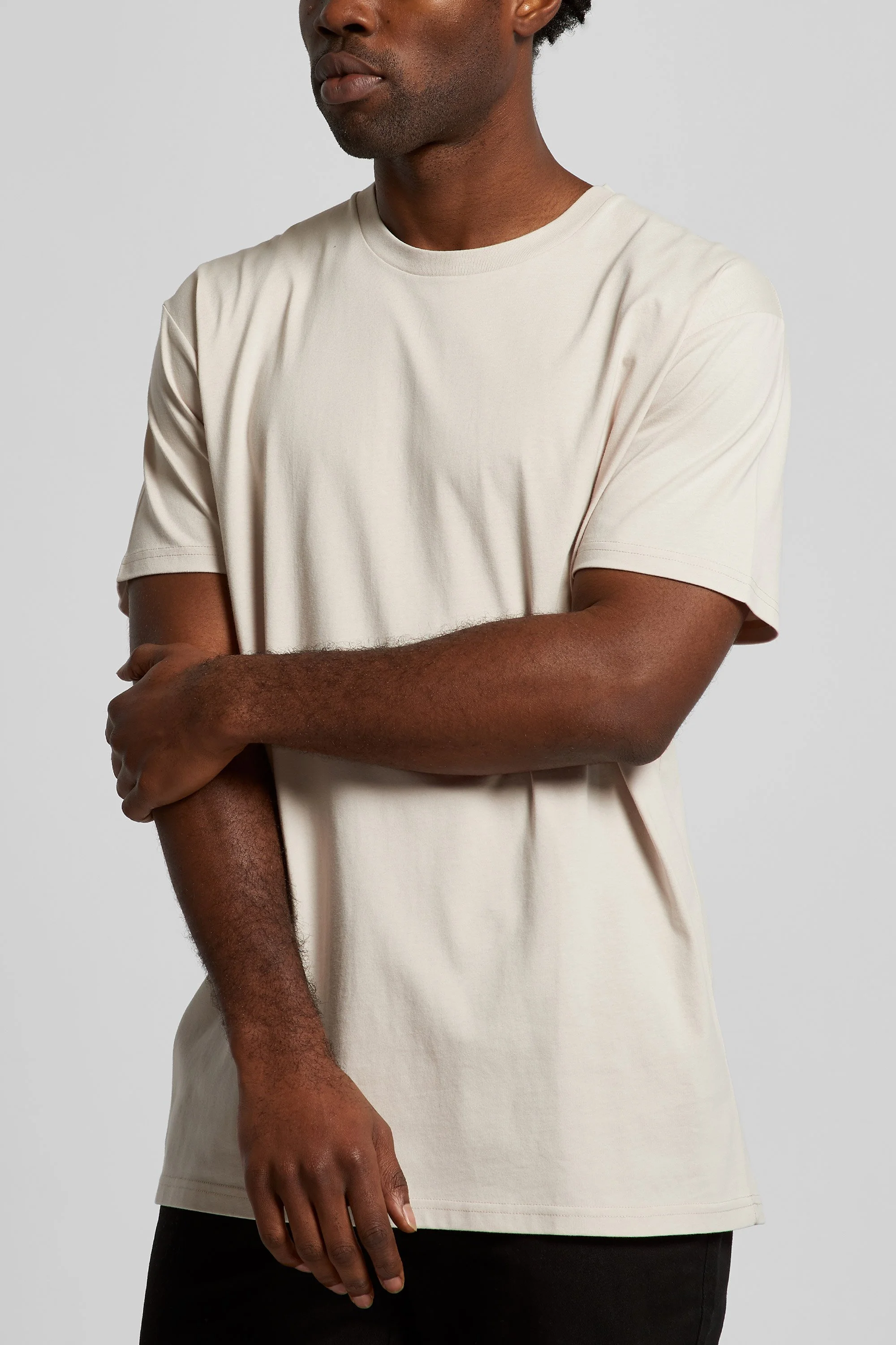 A man with dark skin wearing a plain light beige t-shirt, standing against a light gray background.