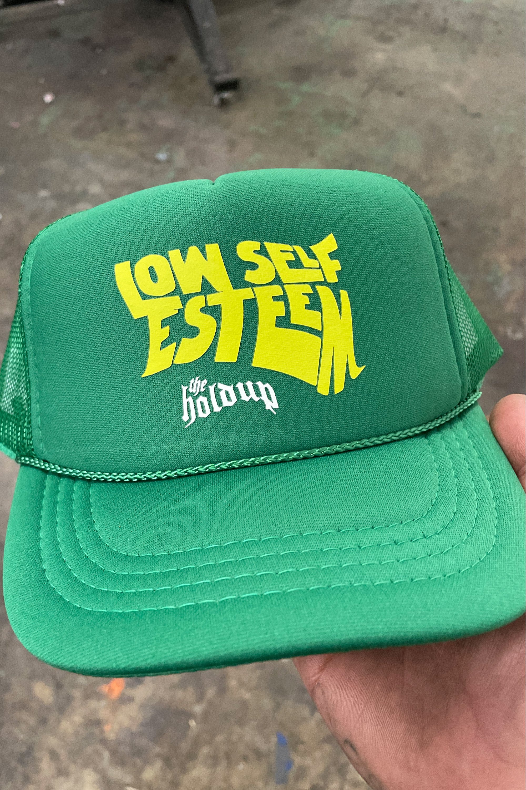 A green trucker hat with yellow and white text that reads 'LOW SELF ESTEEM' and 'the hold up'.