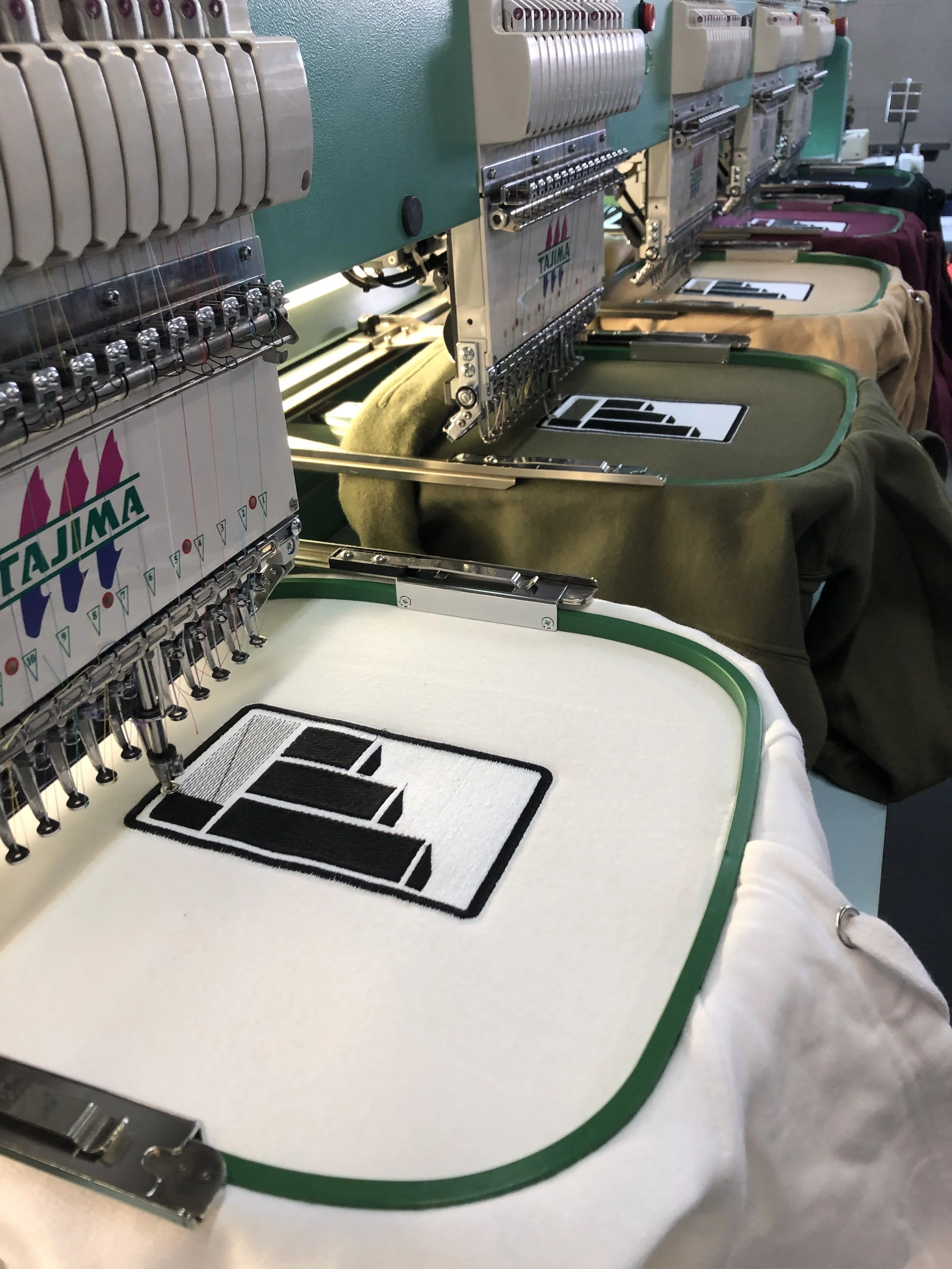 Close-up of embroidery machines stitching a black and white logo onto white fabric with a green border.