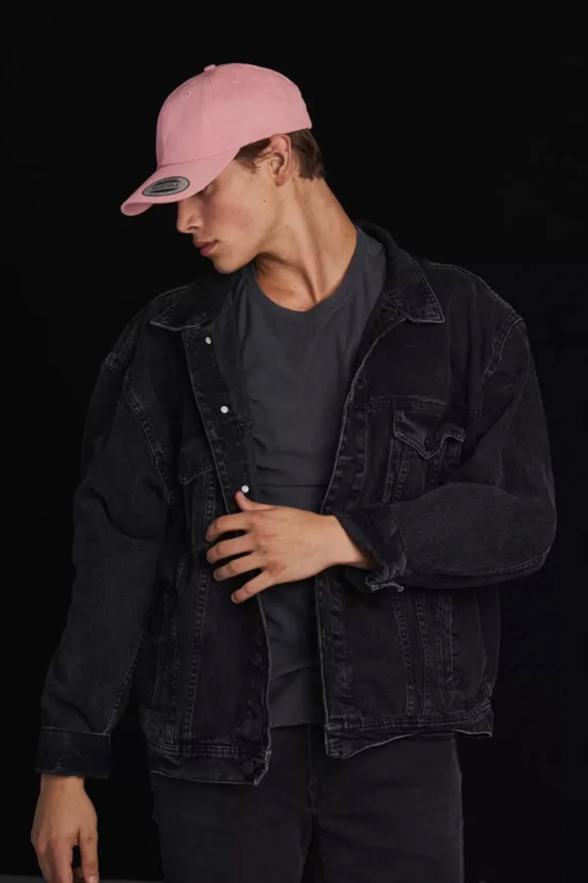 A young man wearing a pink baseball cap, black denim jacket, and dark shirt, standing against a black background, with his head tilted down and eyes closed.