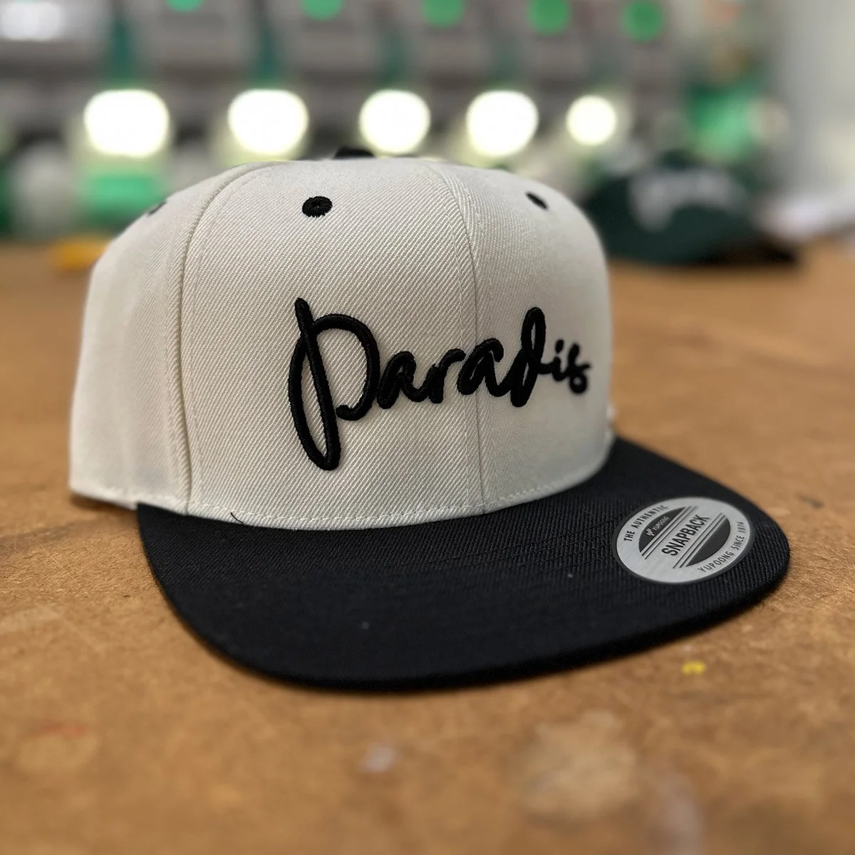 A beige and black snapback cap with the word 'Parafis' embroidered on the front in black script, placed on a wooden surface.