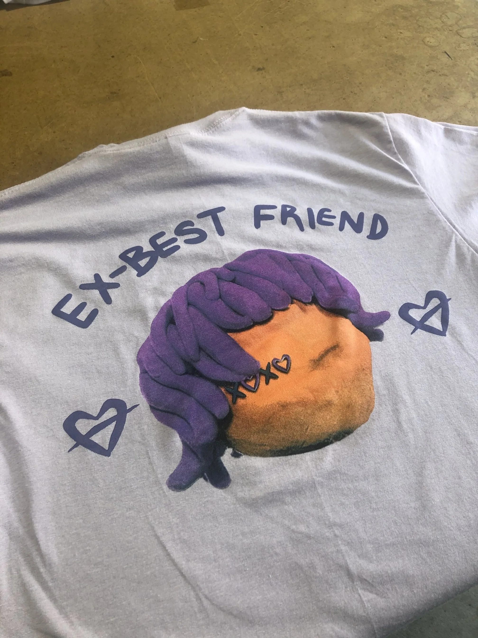 A white t-shirt with a graphic of a peach with purple hair covering one eye, surrounded by the words 'EX-BEST FRIEND' and decorated with small hearts and Xs.