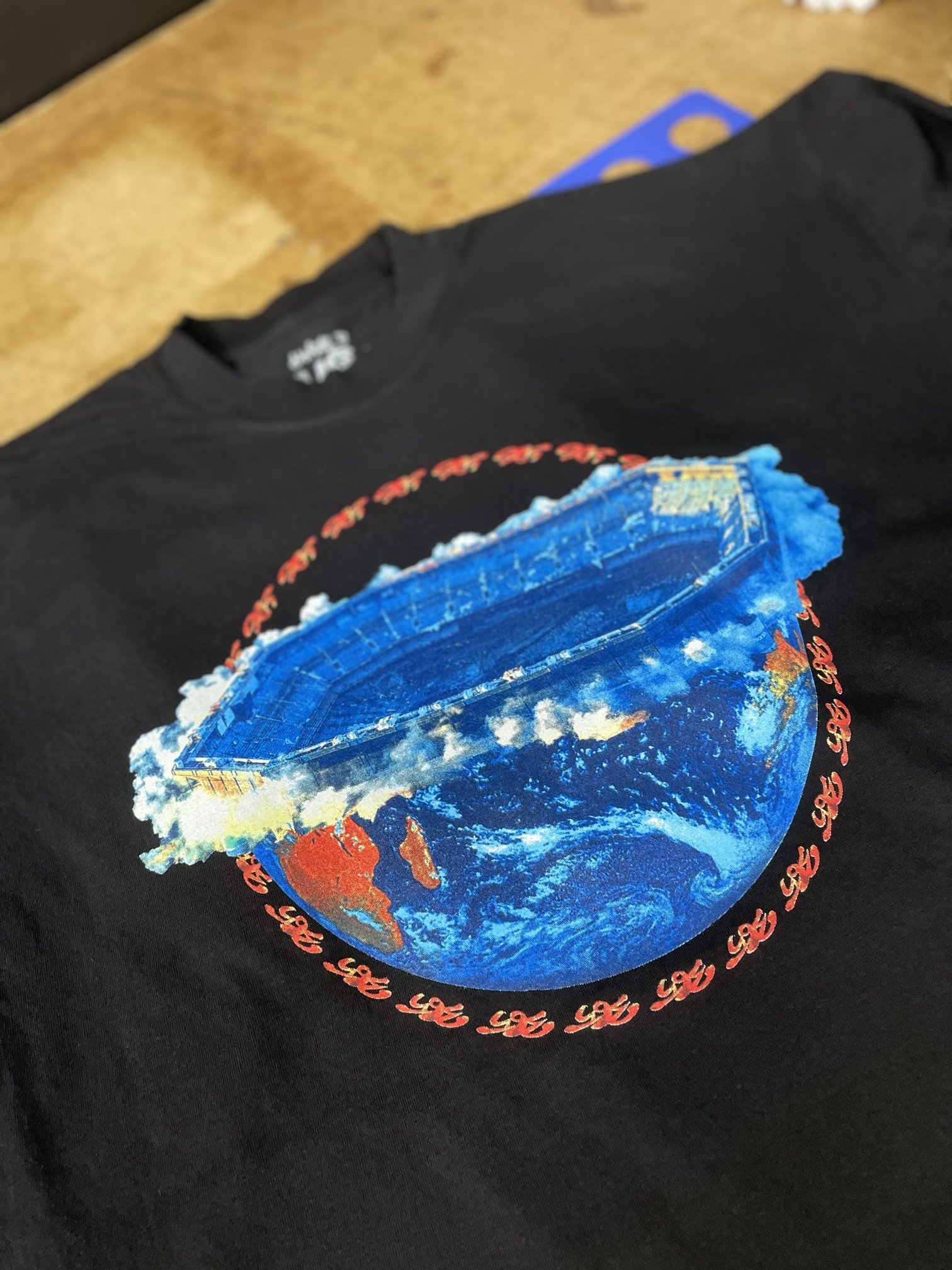 Black T-shirt featuring a graphic of a view of Earth with an upside-down photo of a stadium or amphitheater on top, surrounded by a ring of red cursive text.