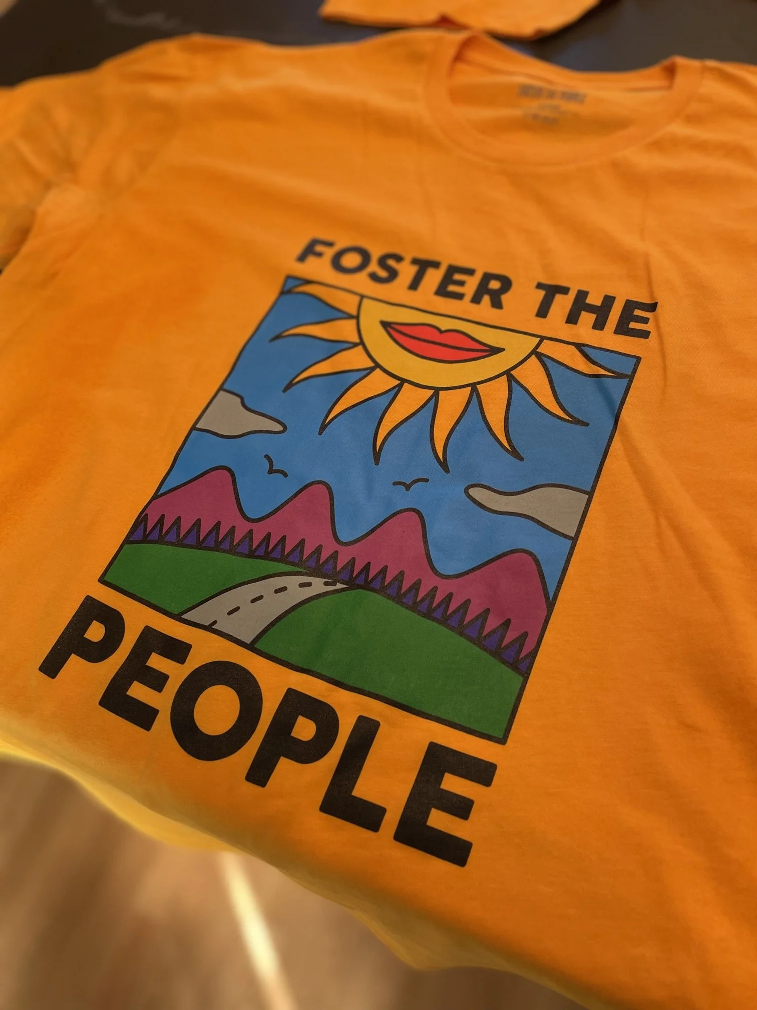 Yellow t-shirt with colorful graphic of a sun, mountains, a road, and birds, with the text 'Foster the People'.