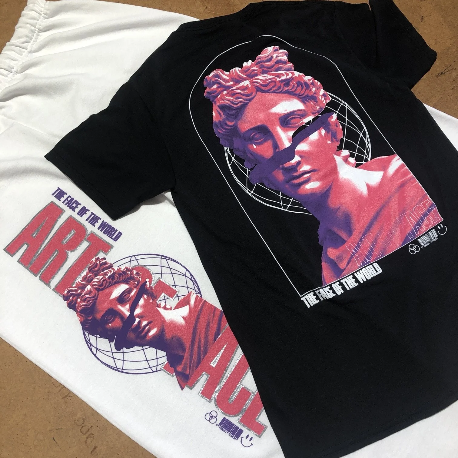Two T-shirts with graphic designs featuring a stylized classical sculpture of a woman's head within a globe outline. The shirts are laid on a flat surface, one white and one black, both displaying similar graphic art and text.