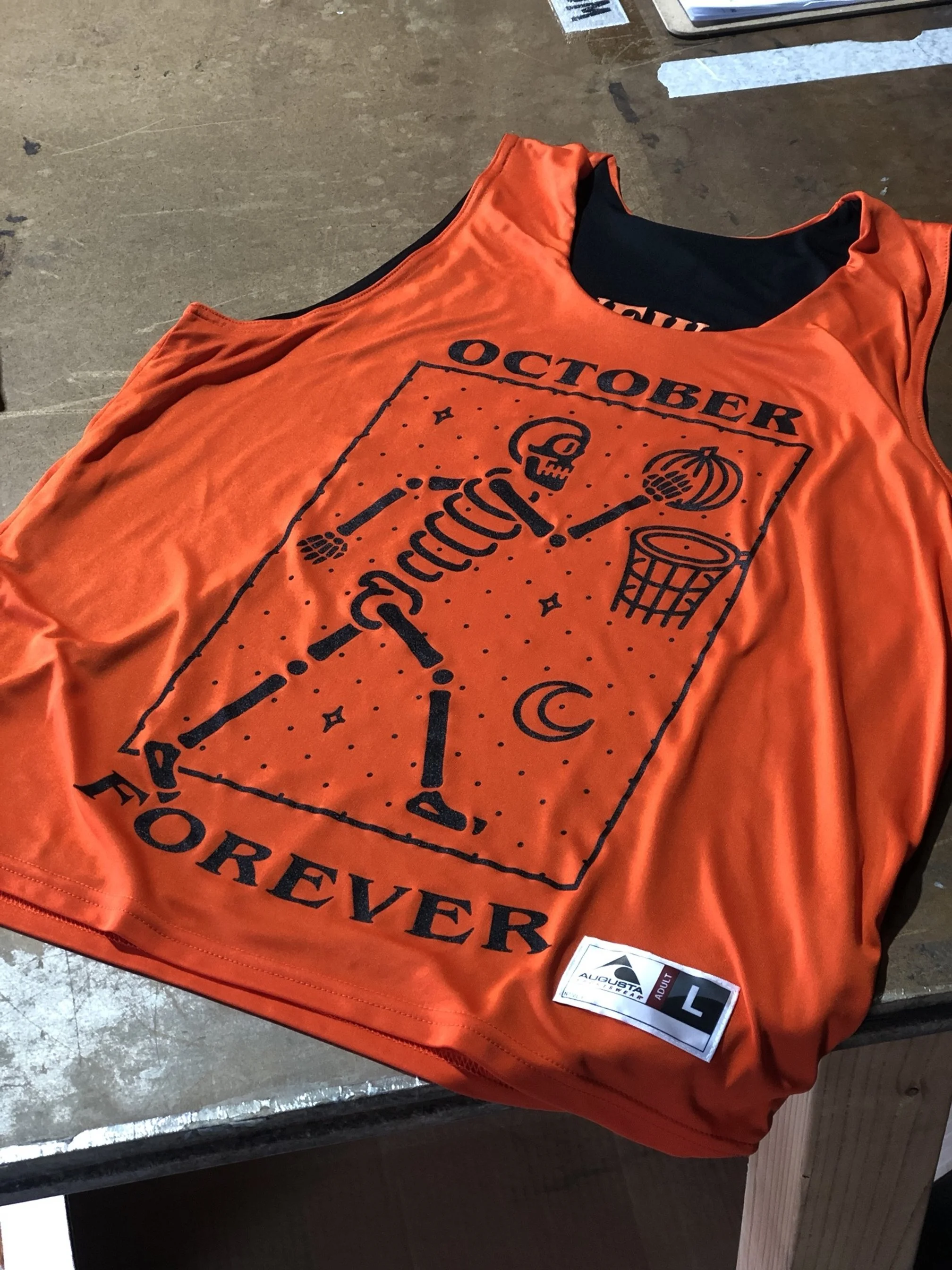Orange sleeveless sports jersey with black printed design showing a golf putter, golf ball, pumpkin, and autumn leaves, with words "October" at the top and "Forever" at the bottom, placed on a wooden surface.