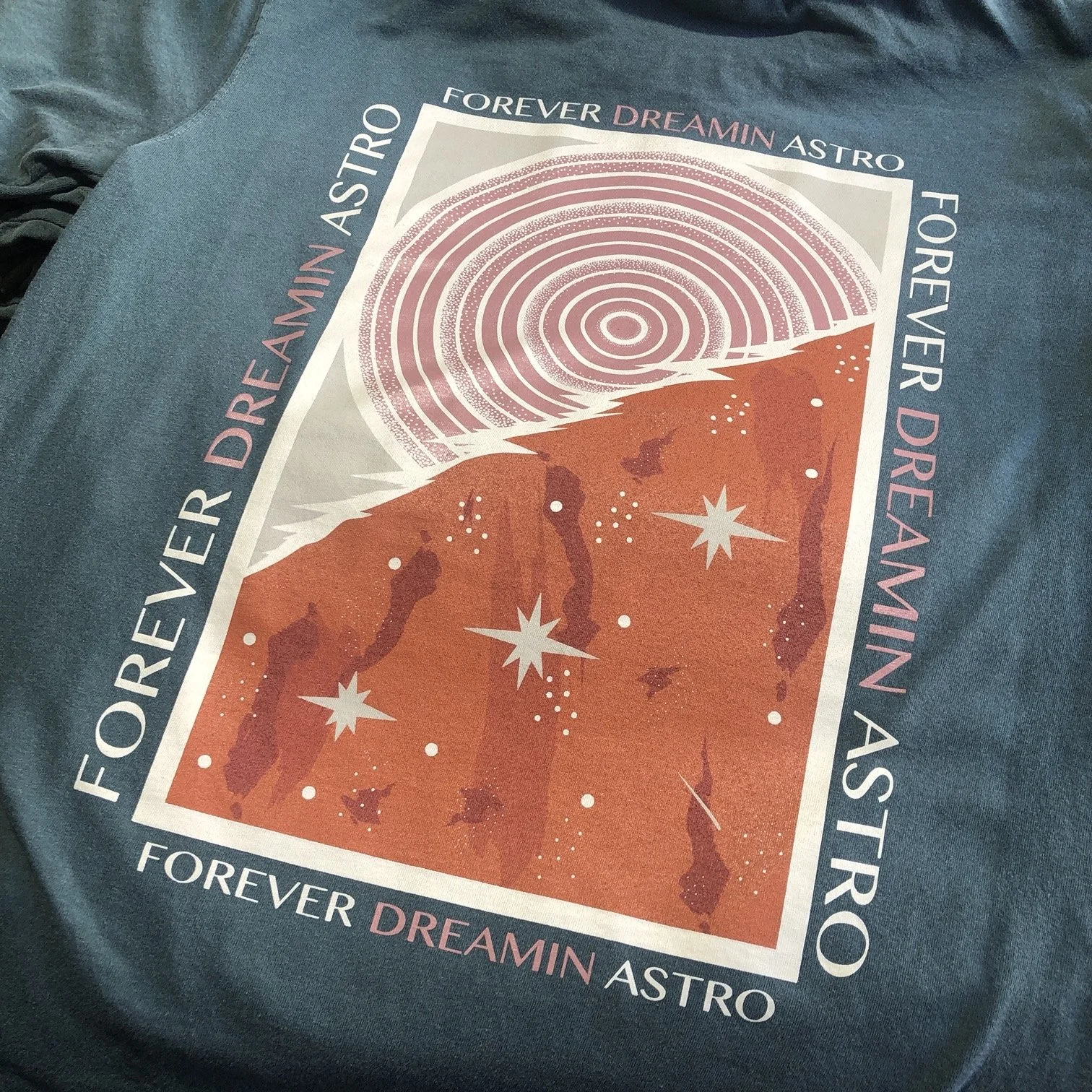 A T-shirt with an astronaut-themed graphic design featuring planets, stars, and swirling sky elements, with text including "Forever Dreamin Astro" and "Forever Dream Astro" arranged around the design.