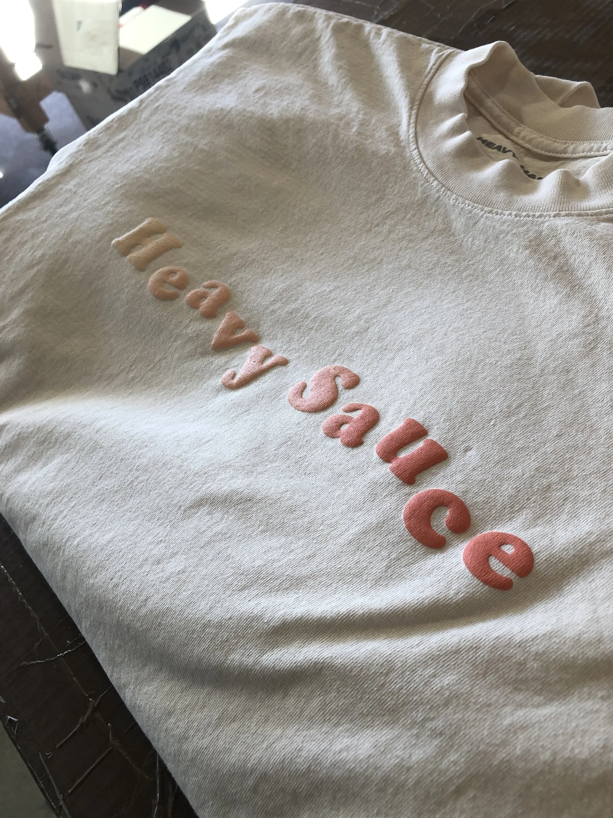 A beige sweatshirt with the words 'Heavy Sailed' embroidered on the front in pastel pink and beige letters, lying on a wooden surface.