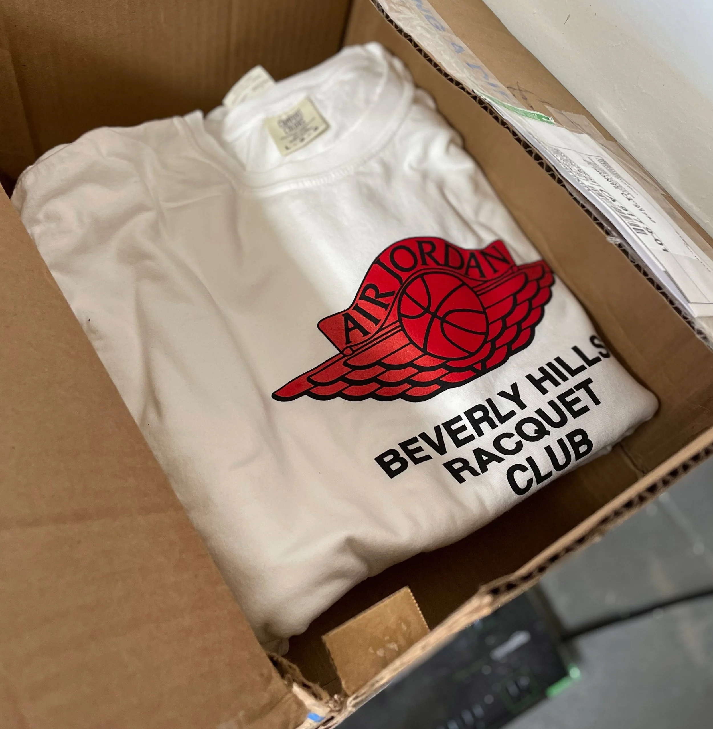 A white T-shirt inside a cardboard box, displaying the logo of the Air Jordan brand with the text 'Beverly Hills Racquet Club' underneath.