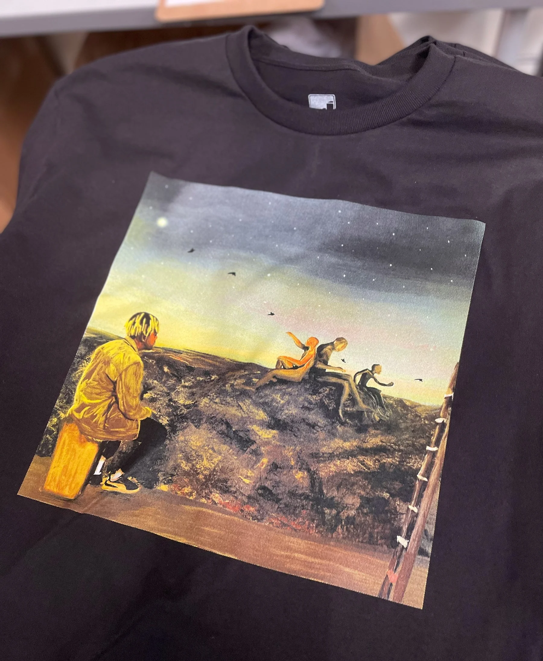 Black T-shirt with a colorful printed scene of people sitting and climbing on a rocky terrain at sunset, with birds flying in the sky.