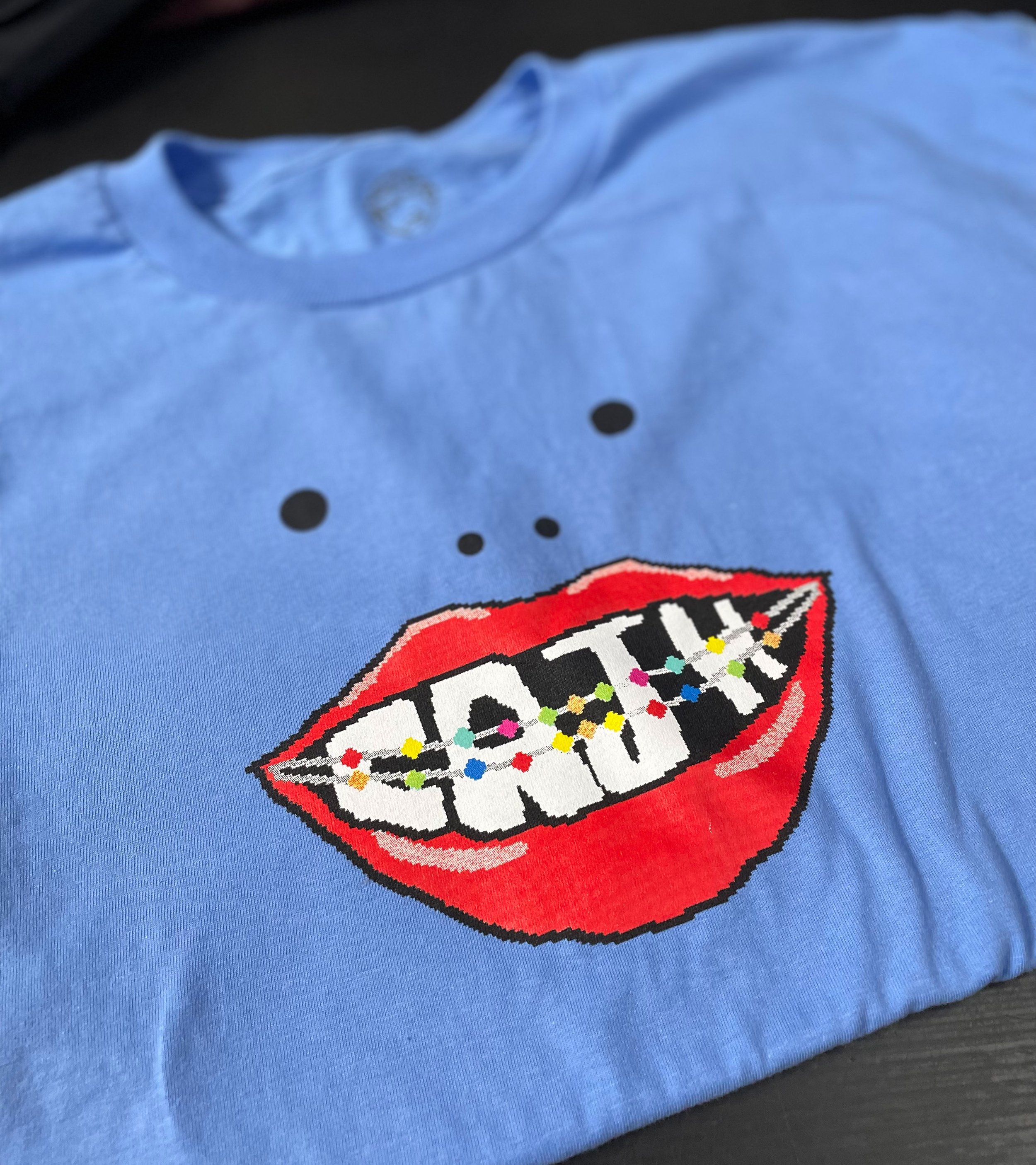 Blue T-shirt with a graphic of open red lips and braces with colorful dots, and black dots above the lips resembling a face.