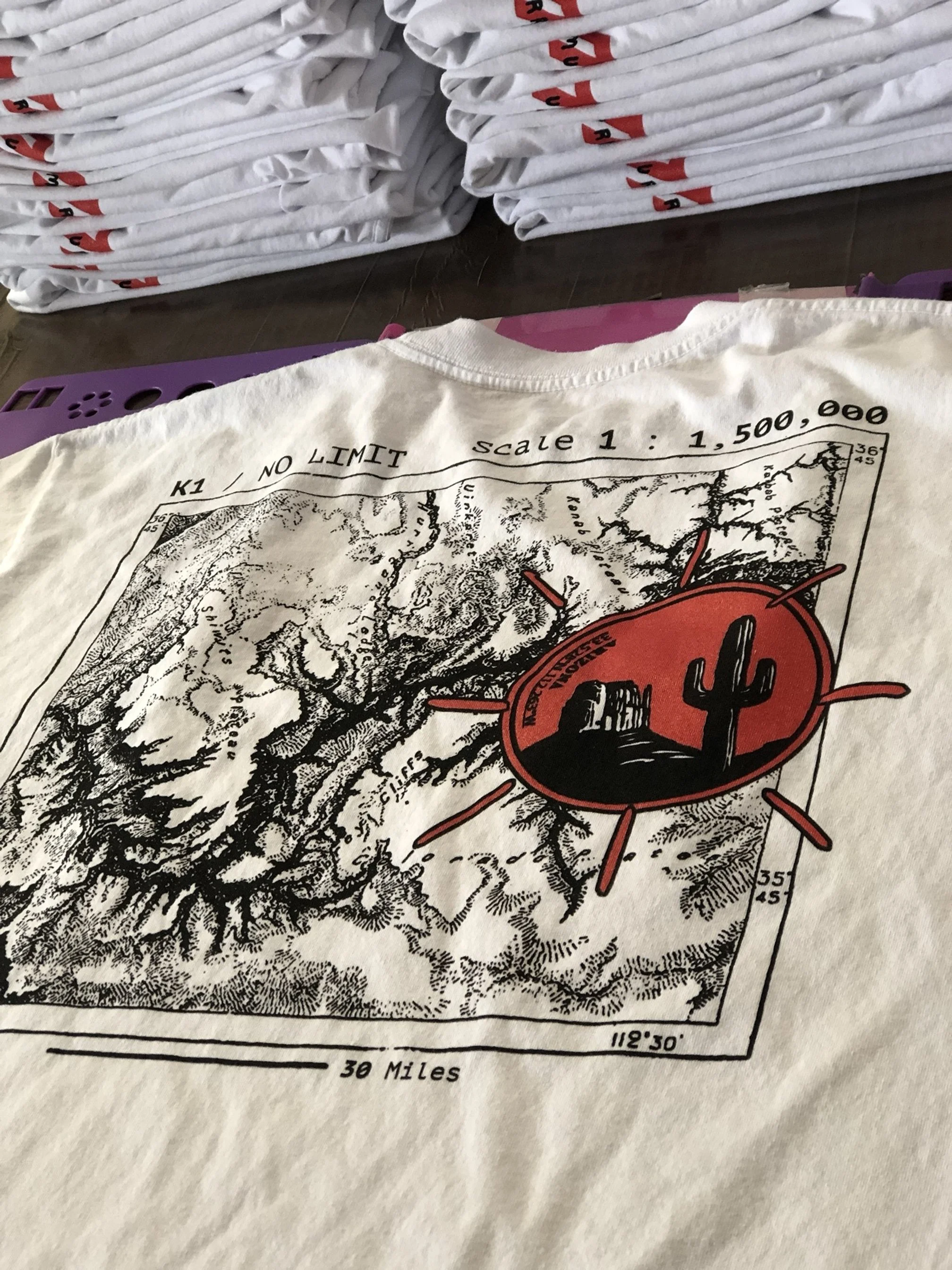 White T-shirt featuring a black and white topographic map of a desert scene with a red cactus emblem in the center, surrounded by red lines.