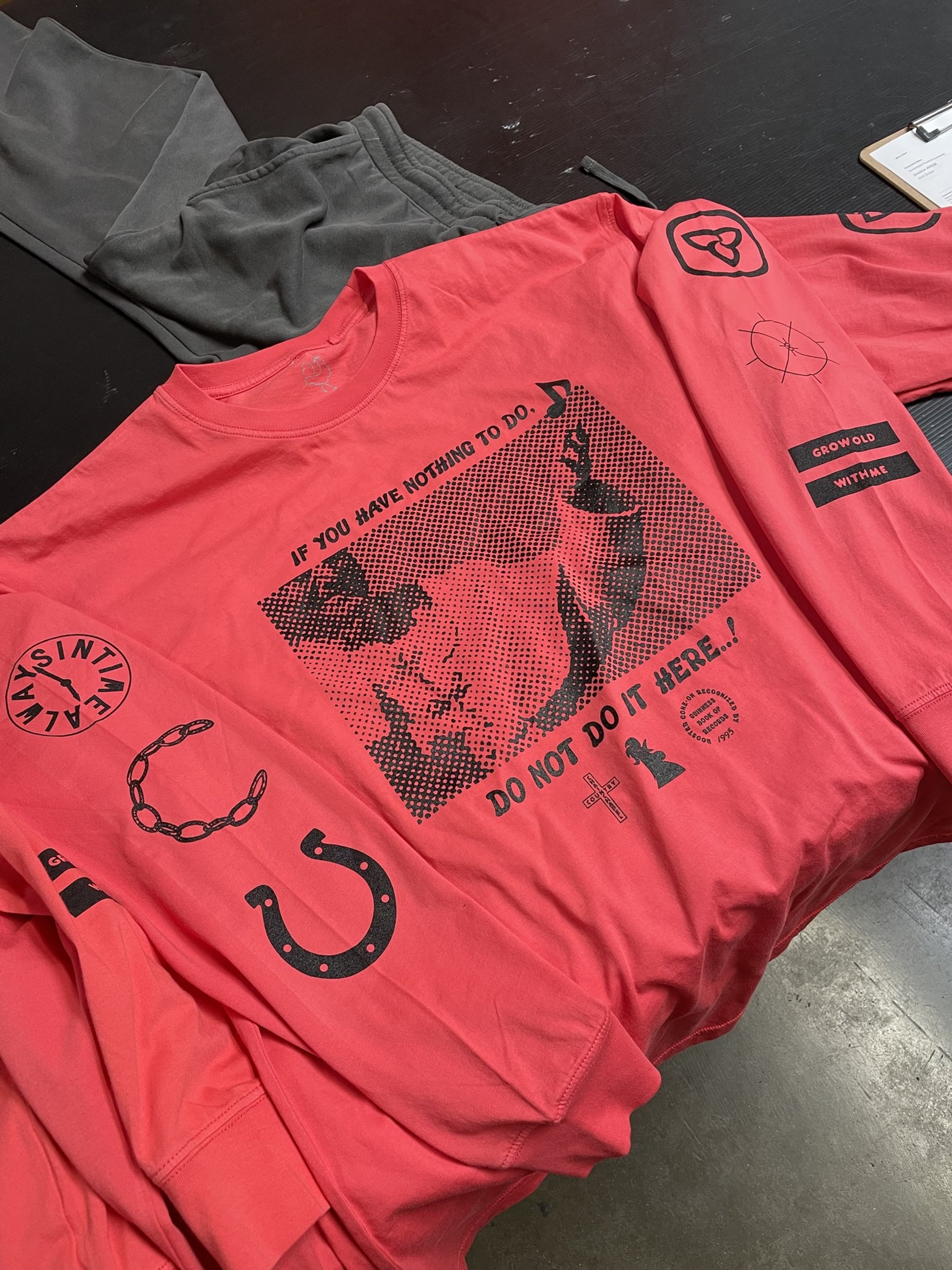 A red T-shirt with black graphic and text, featuring a large portrait of a woman with a hat, surrounded by smaller symbols including a horseshoe, a chain, a compass, and a cross. The text reads, 'If you have nothing to do, do not do it here,' and the