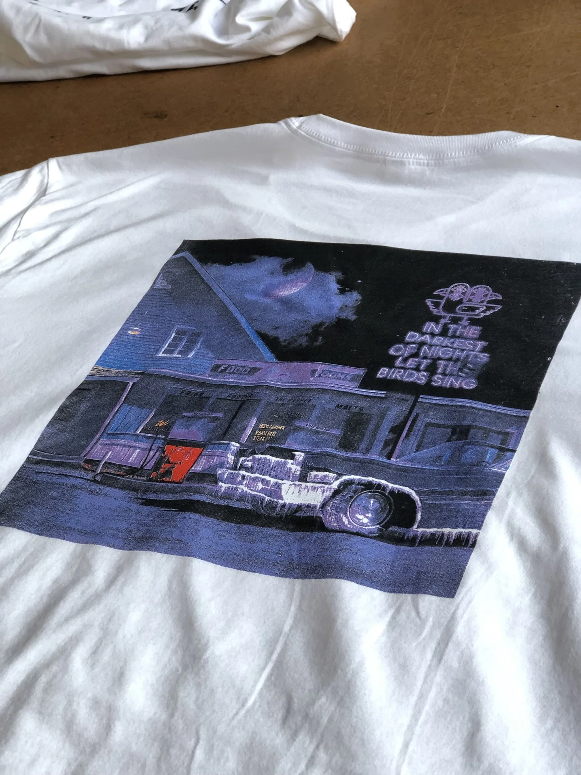 White T-shirt with a graphic print of a vintage car parked in front of a building, with neon-style text and the phrase 'In the darkest of nights, let the birds sing'.