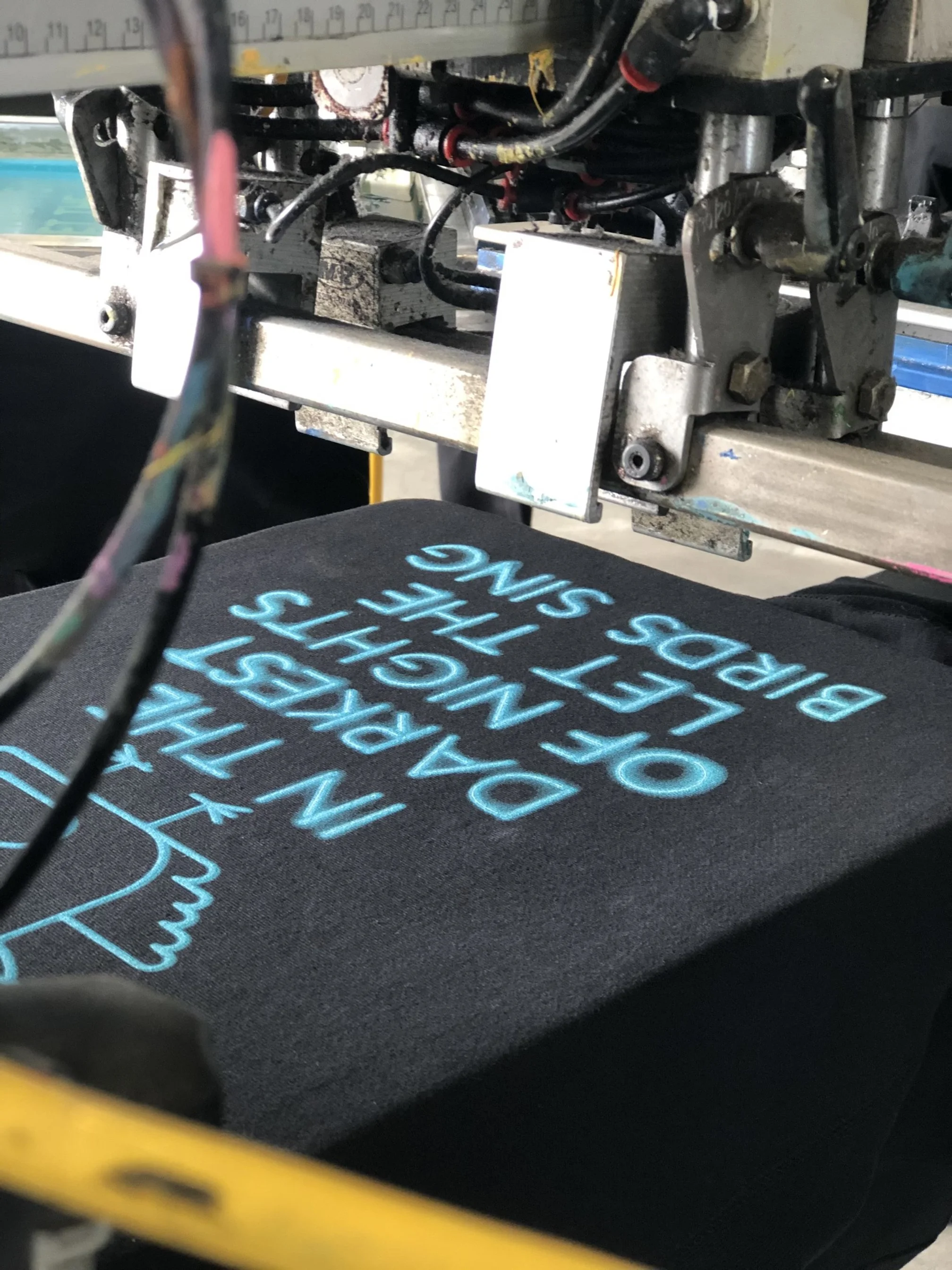 Close-up of embroidery machine stitching light blue text onto a black fabric, with the machine's metal and wiring visible above.