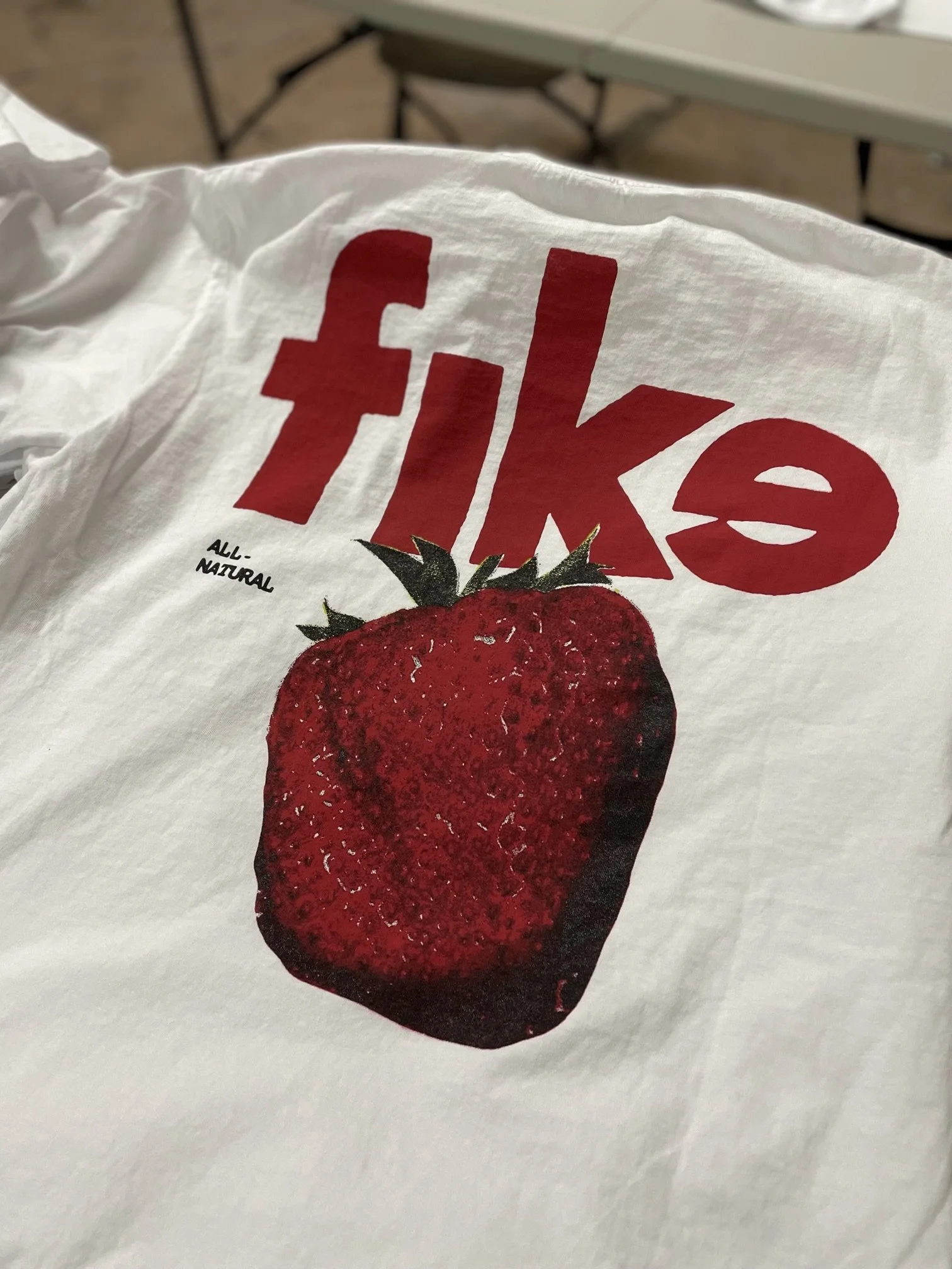 Close-up of a white t-shirt featuring the word 'Fiji' in red capital letters with a stylized 'i' filled with a red circle, and an image of a large red strawberry with green leaves, labeled 'All-Natural'.