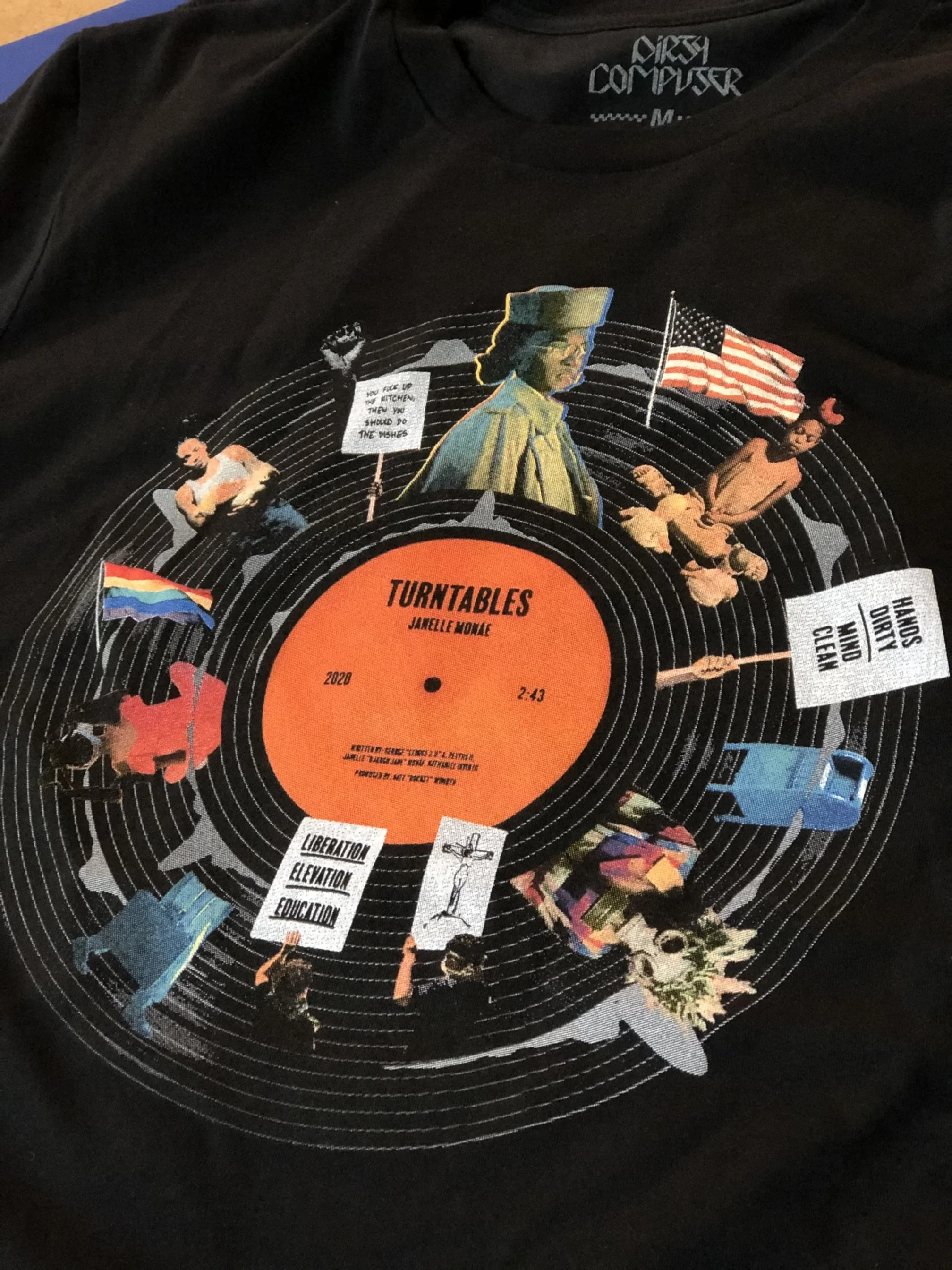 A black T-shirt with a design featuring an orange vinyl record labeled "Turntables" by Janelle Monae, with a collage of images including a woman in a military-style hat, people holding protest signs, and flags, set against concentric circles.