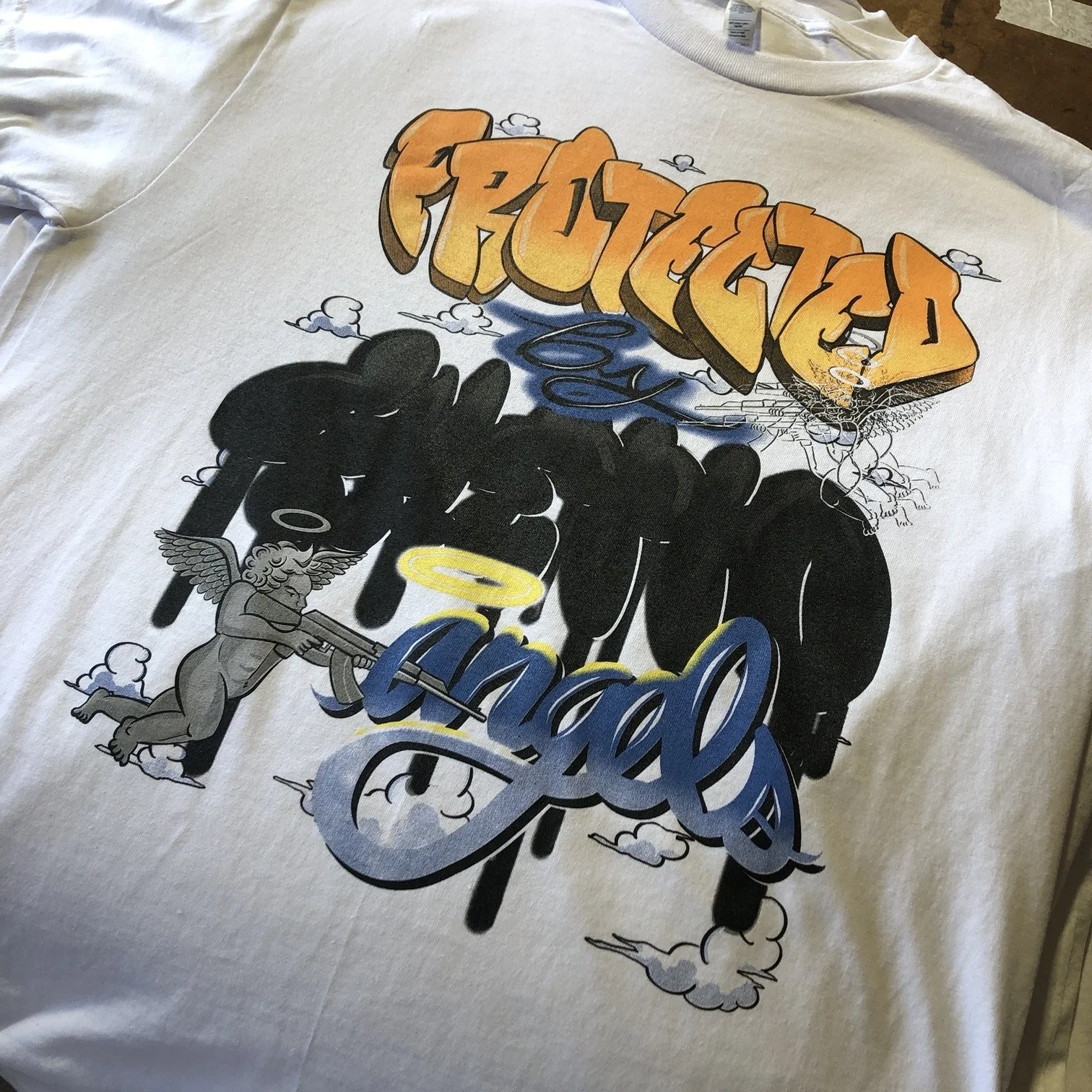 A white T-shirt with graffiti-style text that reads "Fugitives" in orange and yellow letters, with additional words "Intern" in black, and "Angel" in blue. The design includes a cherub holding a rifle, a yellow halo, and cloud-like elements.