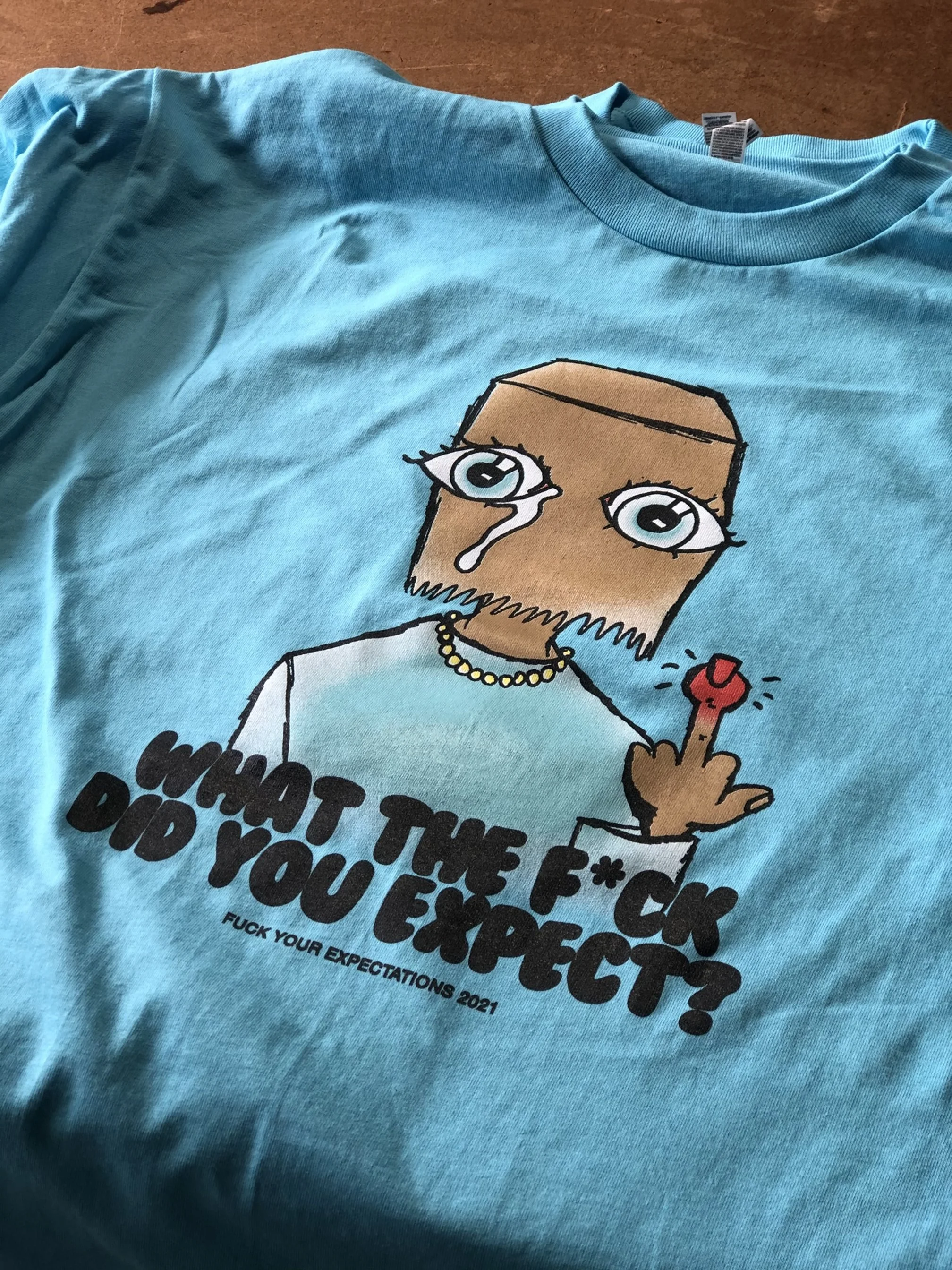 A blue T-shirt featuring a cartoon character with a cardboard box for a head, crying with tears and holding up a middle finger. The shirt has text that says, "WHAT THE F**K DO YOU EXPECT?" and "F*CK YOUR EXPECTATIONS 2021."