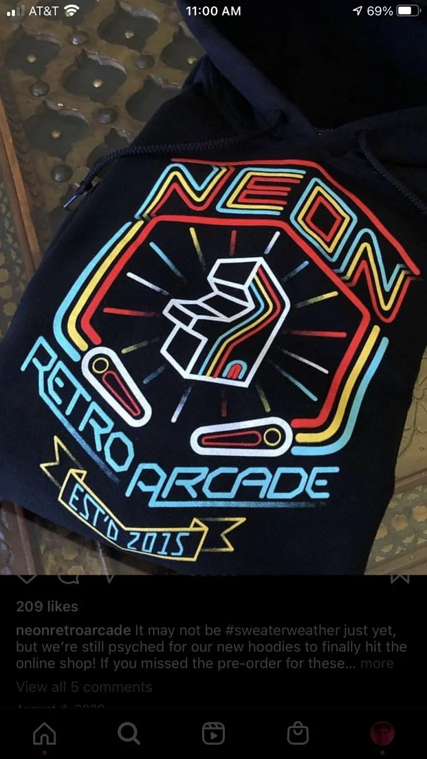 Close-up of a black hoodie with neon-colored graphics and text. The design features a vintage arcade game style with the words 'NEON', 'RETRO ARCADE', and 'ESTD 2015'.