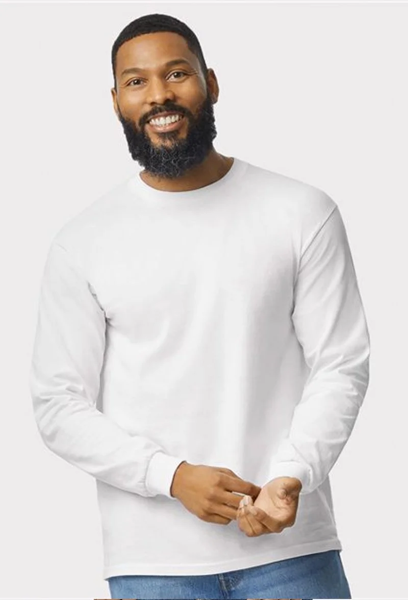 A man with dark skin, a beard, and short hair smiling, wearing a white long-sleeve shirt and blue jeans, standing against a plain light gray background.