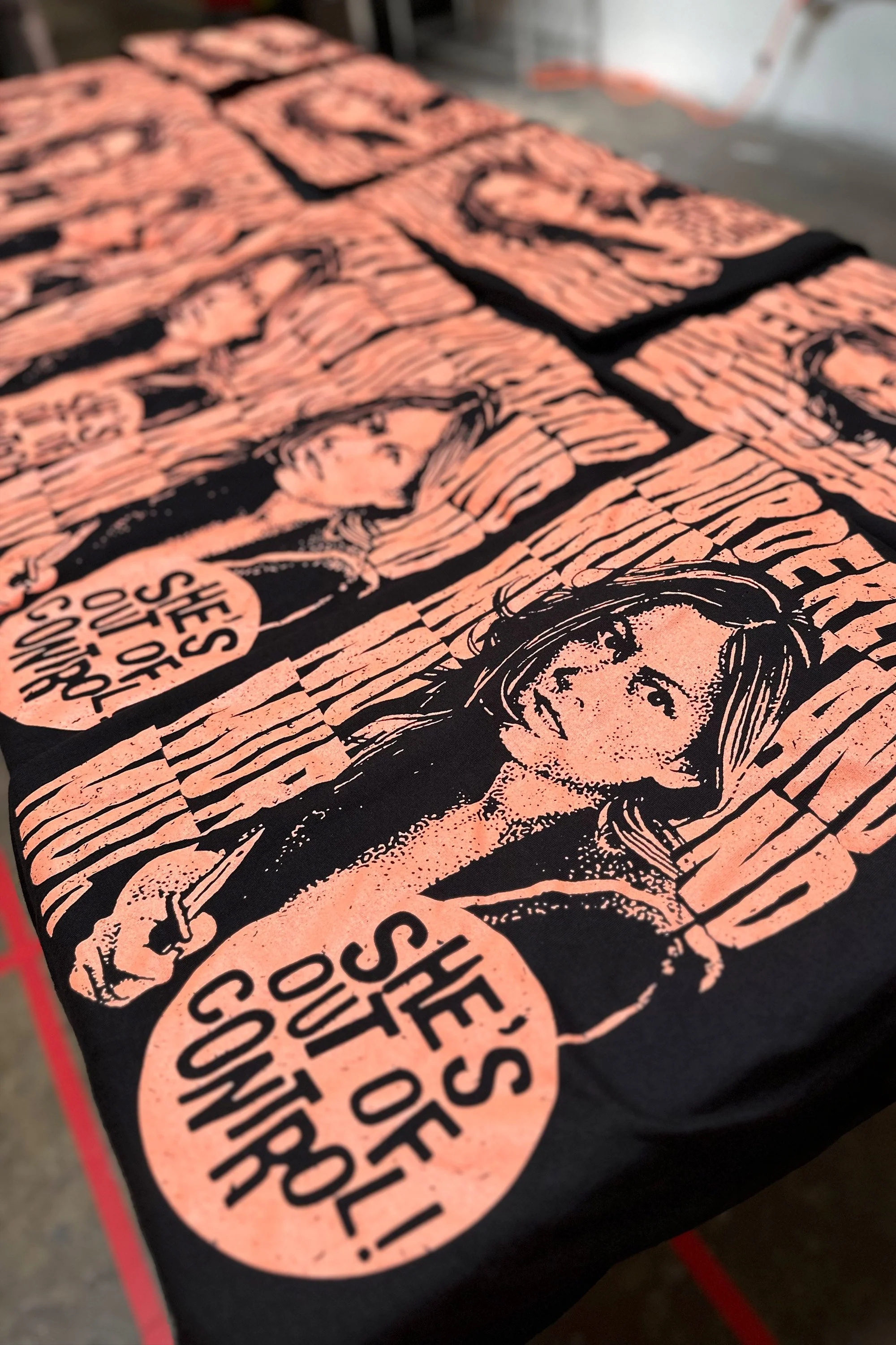 Close-up of a black T-shirt with a pink graphic of a woman holding a sign that says 'She’s out of control!'.