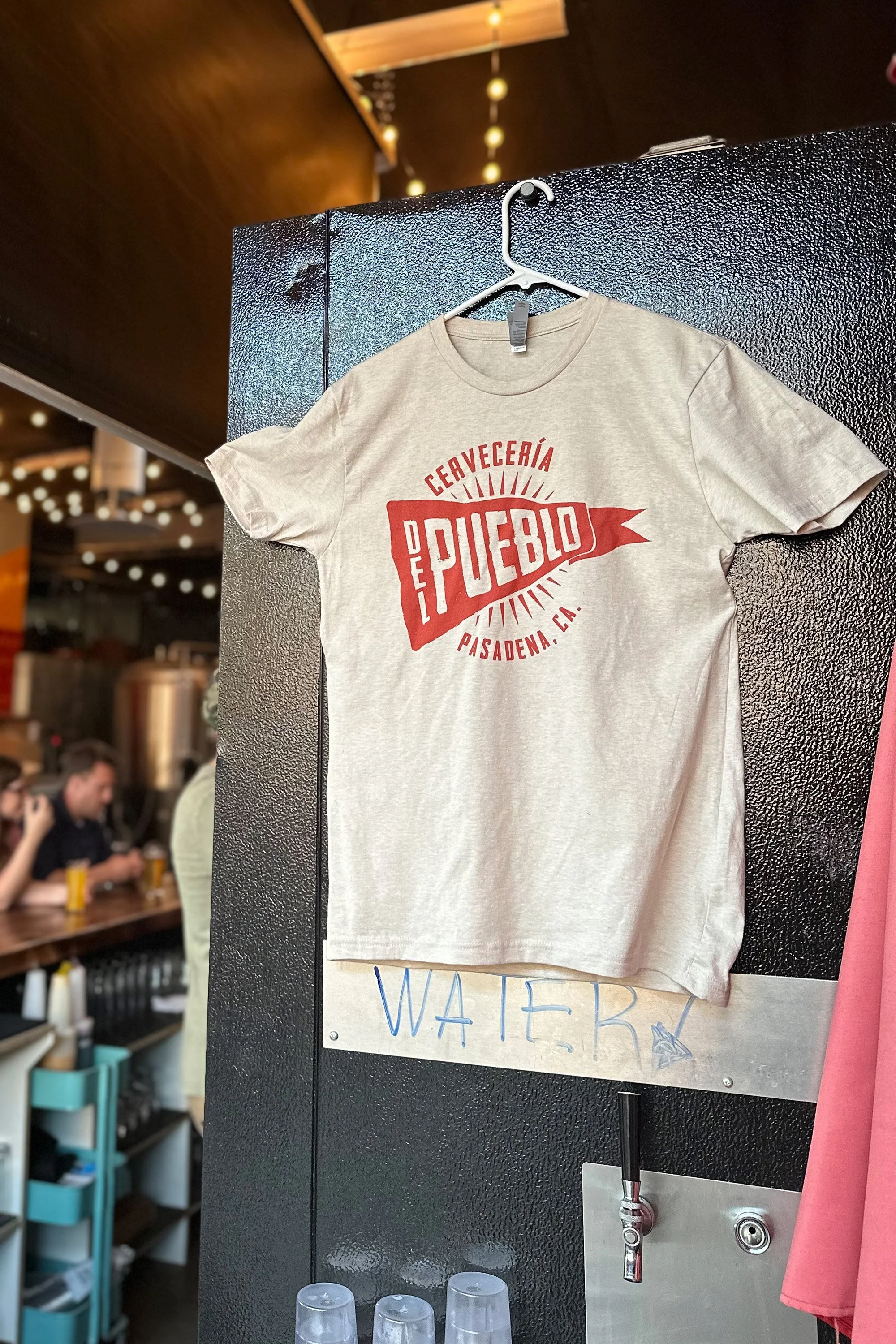 A beige T-shirt hanging on a hook with red text that reads protuberantly 'CERVECERÍA DEL PUEBLO PASADENA, CA.' The background shows a part of a bar or restaurant with people and lighting.