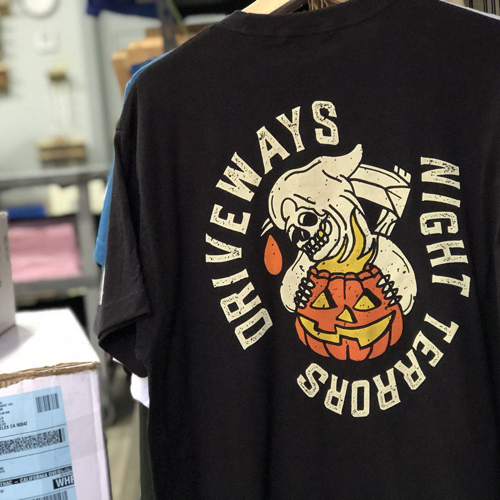 A black T-shirt with a Halloween-themed graphic of a skull, a drooping orange tear, and a pumpkin with a carved face. The text surrounding the graphic reads "DO YOU ALWAYS NIGHT LIGHT YOURS" in a circular pattern.