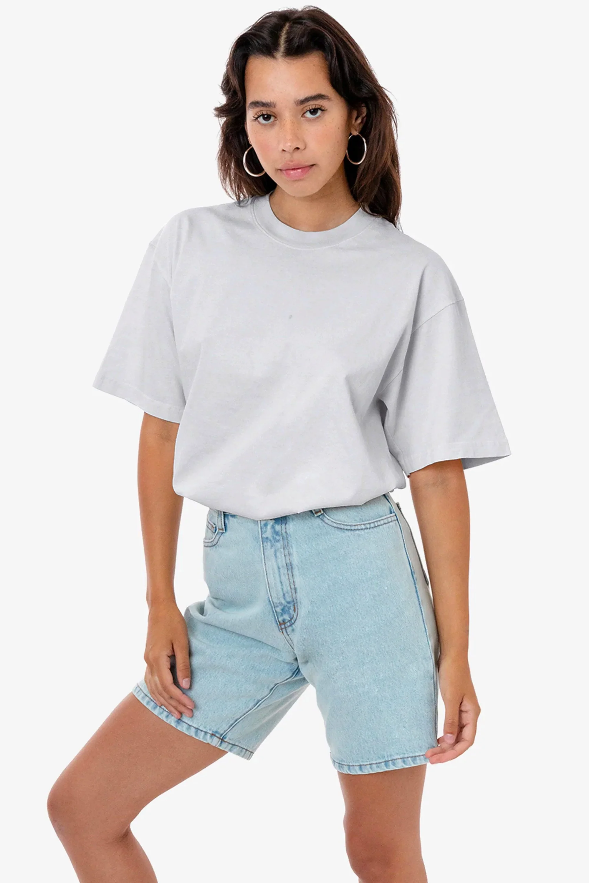 A young woman with dark hair, wearing hoop earrings, a gray t-shirt, and light-wash denim shorts, standing against a plain white background.