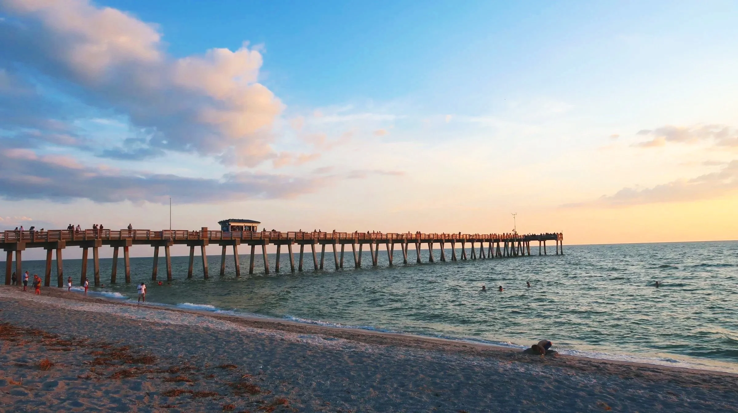 A wooden pier extending into the ocean at sunset with people walking and swimming nearby, partly cloudy sky with soft pastel colors.